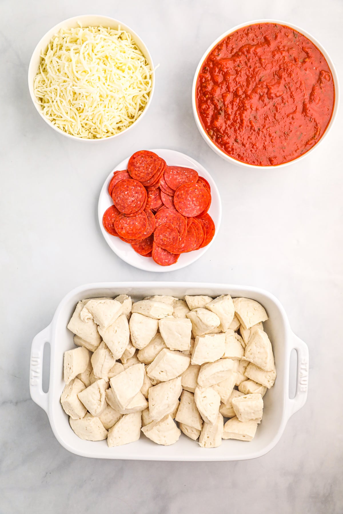 a casserole dish with bits of biscuit dough next to three bowls with pizza sauce, cheese, and pepperoni