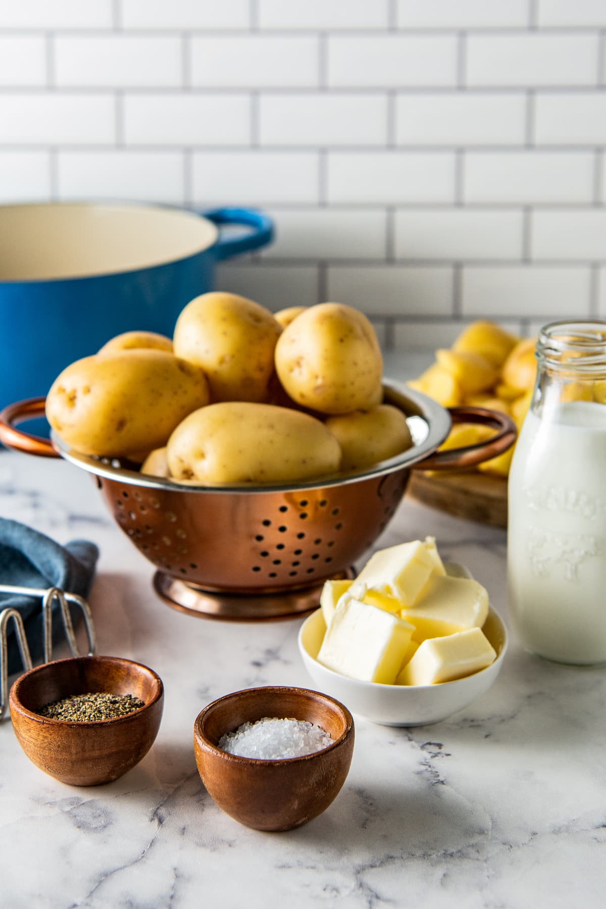 a colander with whole potatoes on the counter with a container of cream, bowl of butter, salt and pepper