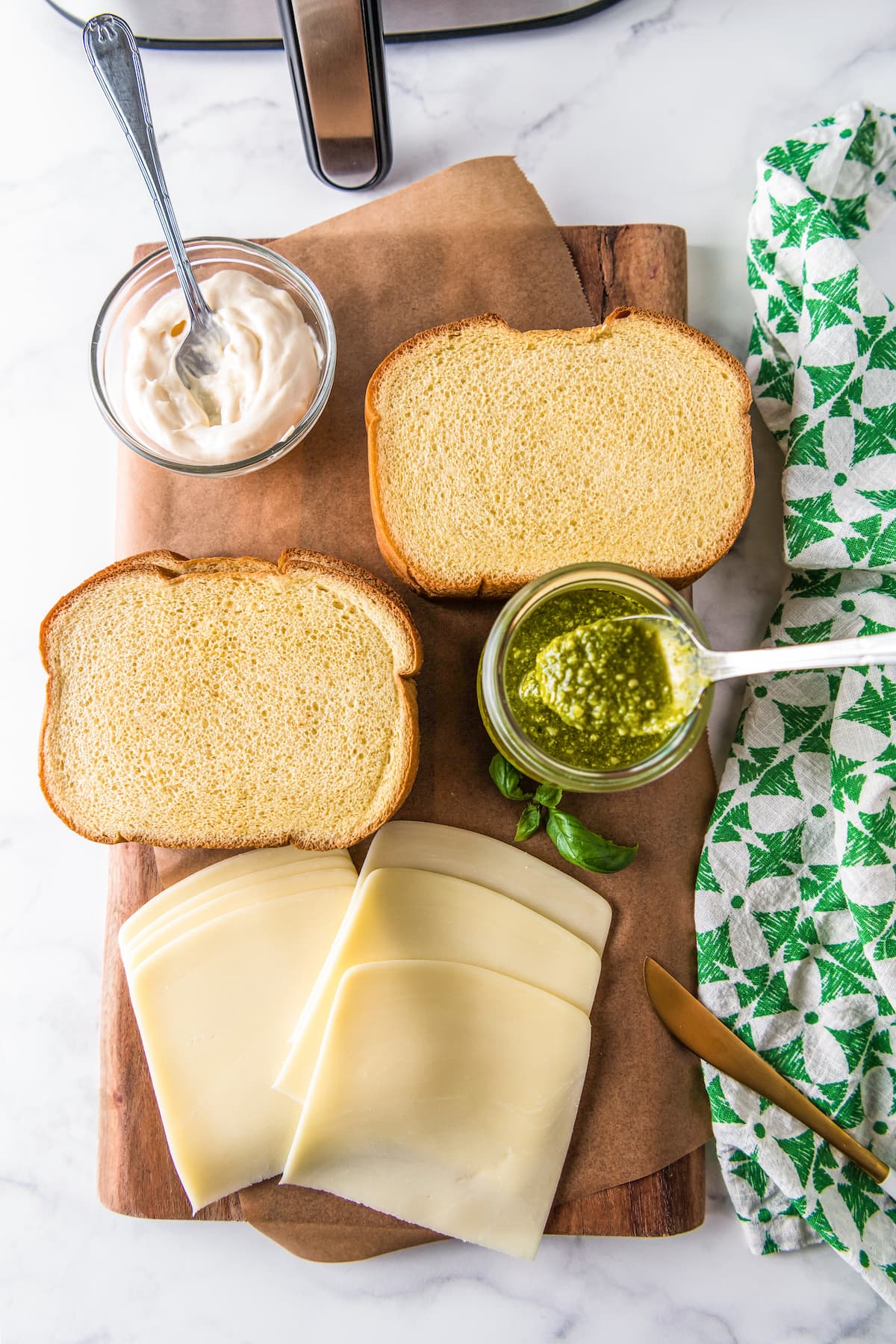 a cutting board with mayo, pesto, sliced cheese, and two slices of bread
