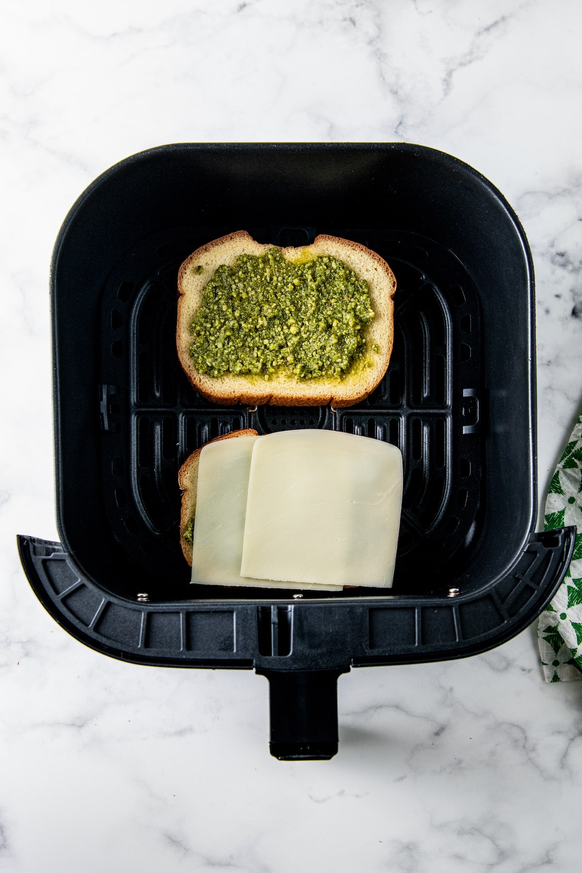 two slices of bread in an air fryer tray, one with pesto and one with cheese on top