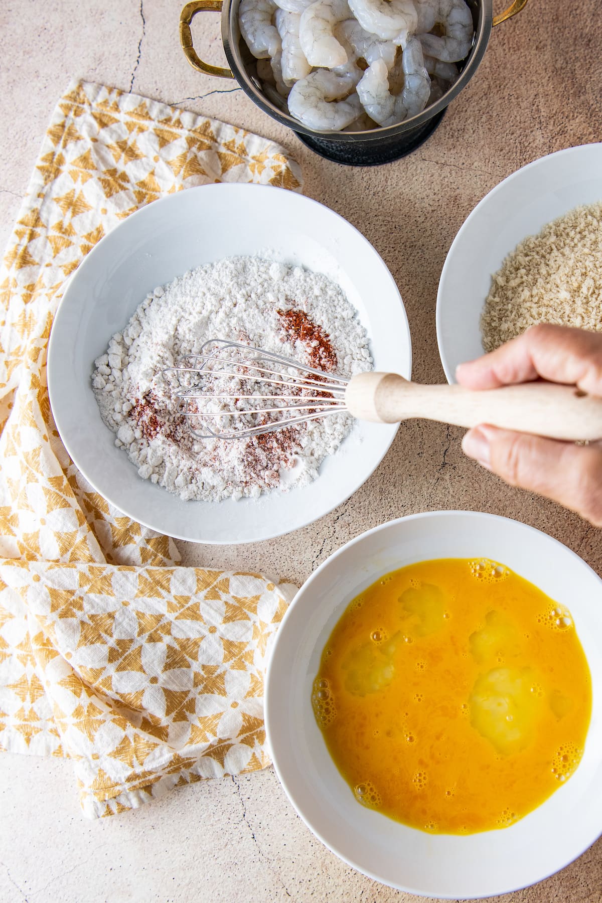 a bowl with flour and spices being whisked together next to bowls of panko and mixed eggs