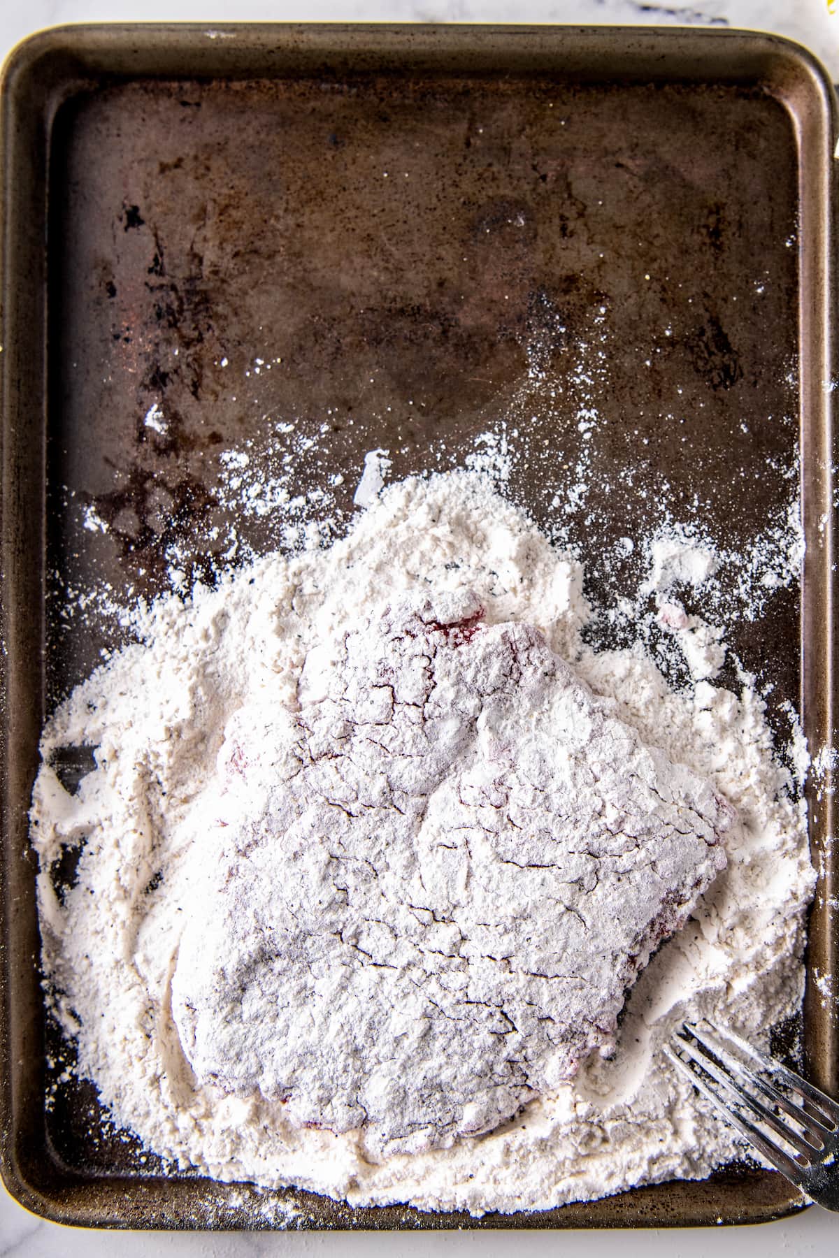 cubed steak being coated with flour on a sheet tray