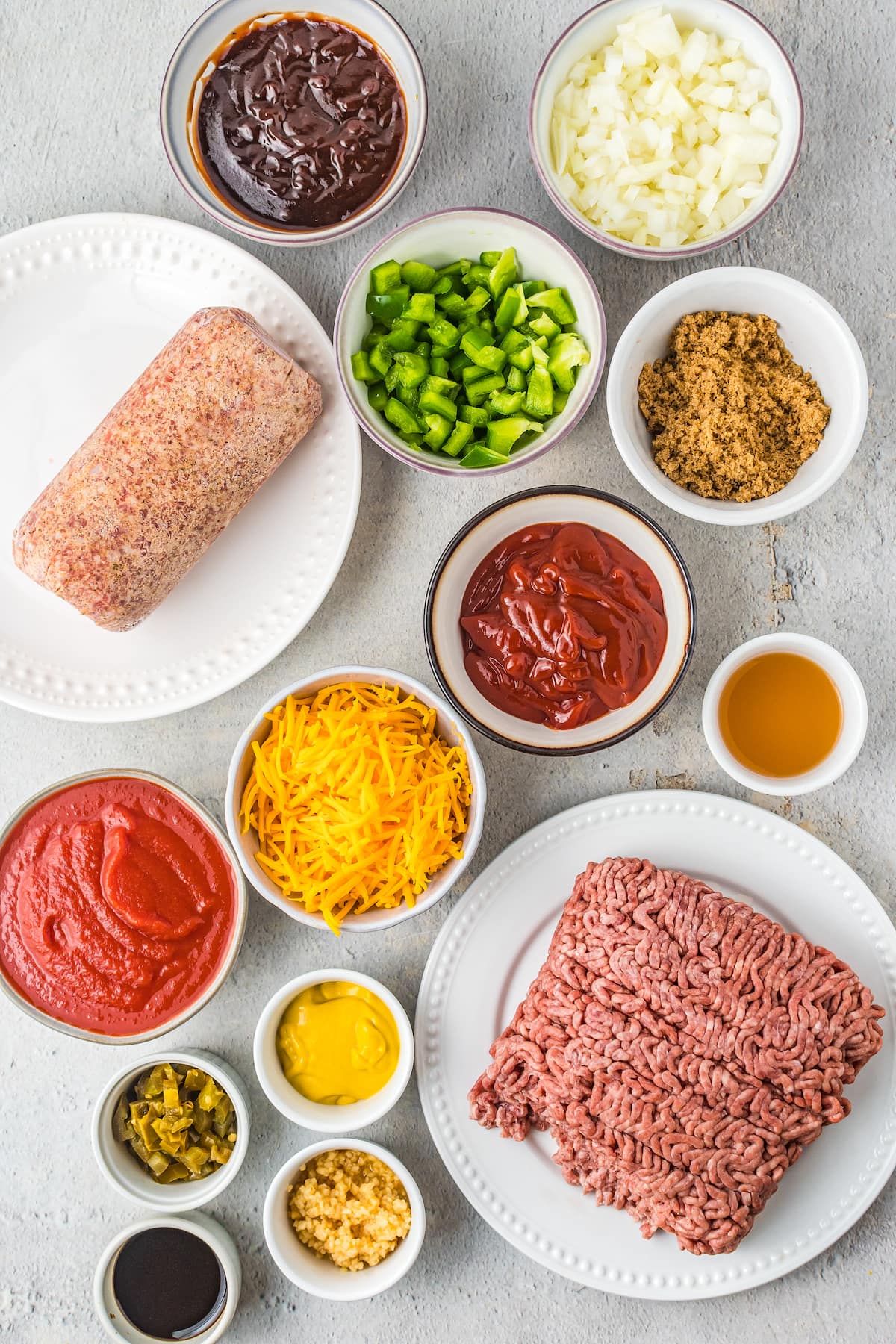Ingredients to make sloppy joes like ground beef, ground sausage, ketchup, bbq sauce, tomato sauce, chopped peppers, chopped onions, and shredded cheese