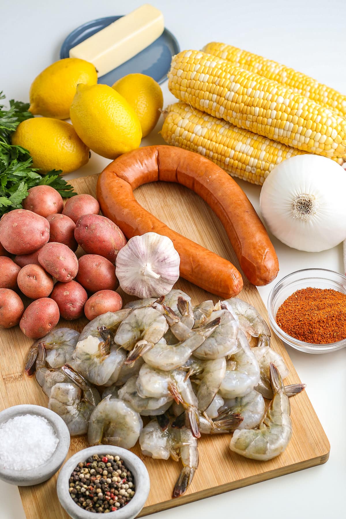 cutting board with raw shrinmp, potatoes, garlic, sausage and corn on the cob, lemons, one onion and seasoning on the side