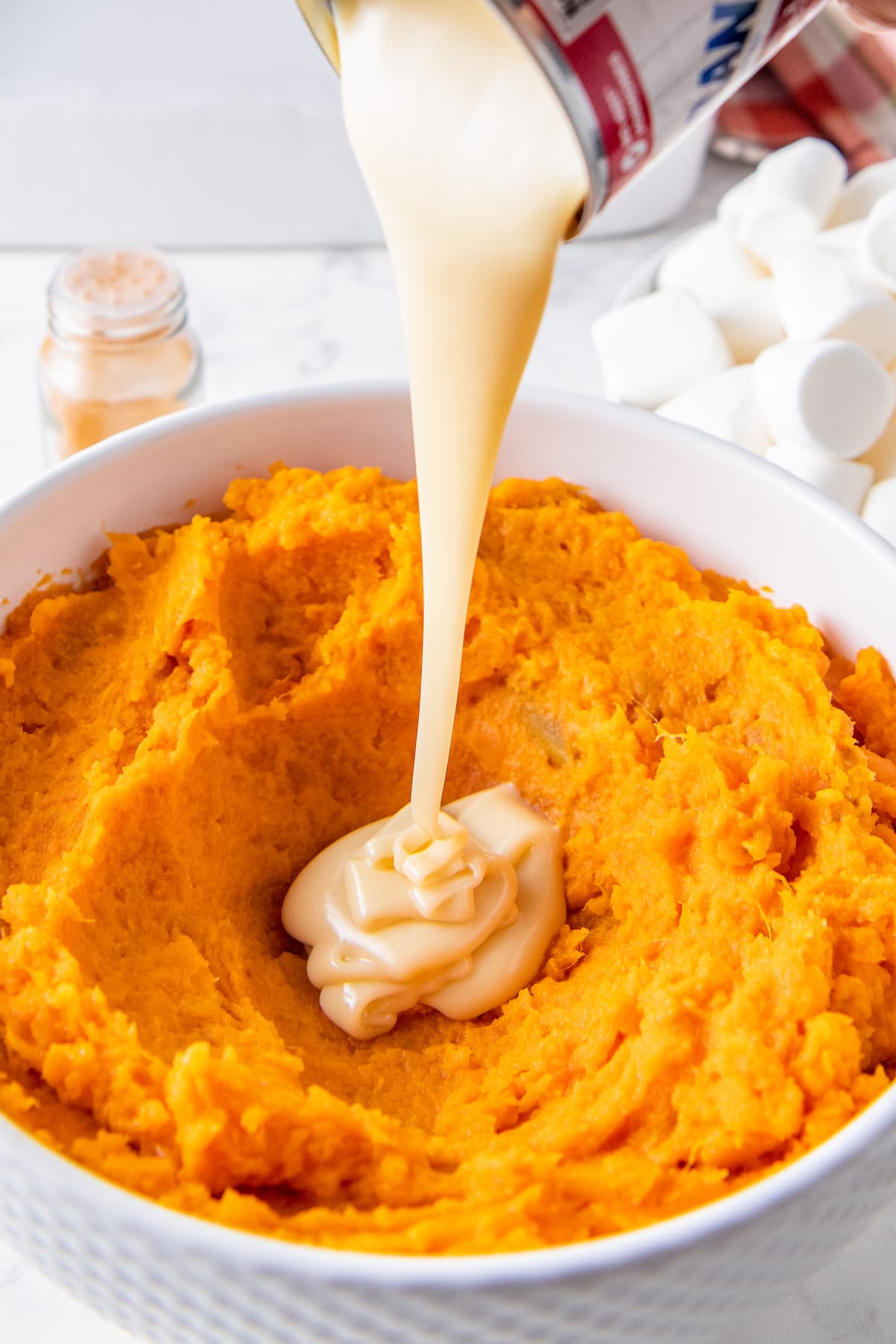 pouring sweetened condensed milk into a bowl of mashed sweet potatoes