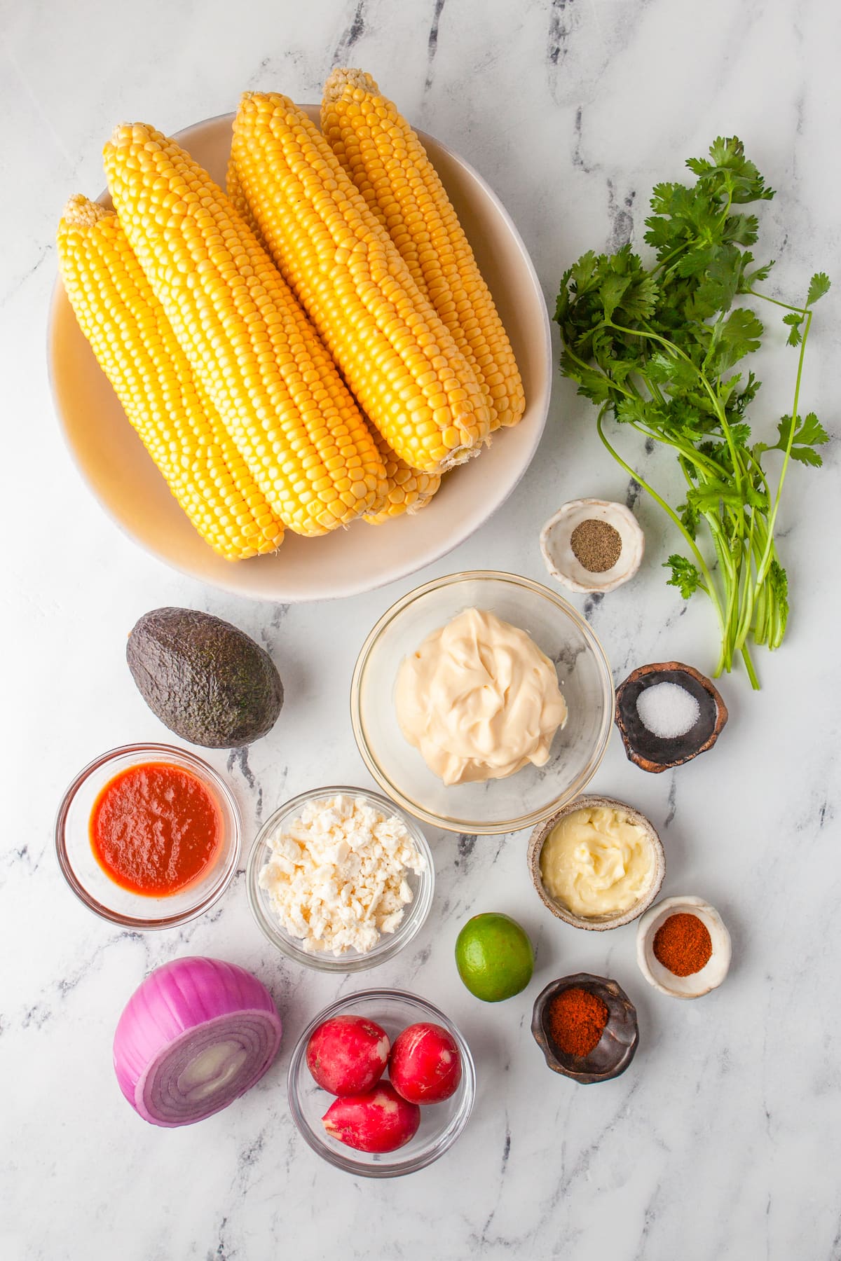 a tablescape of corn on the cob, fresh cilantro, mayonnaise, cotija cheese, avocado, chili powder, salt, and pepper