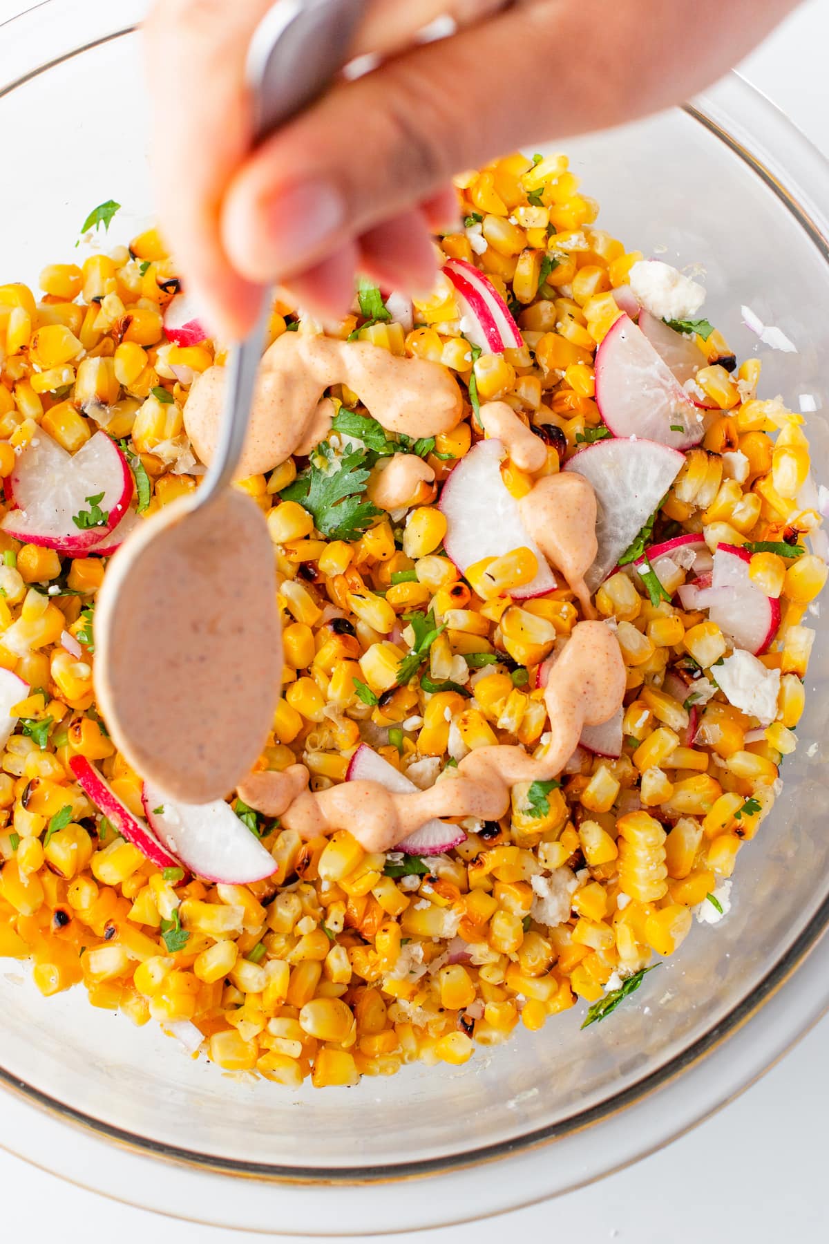 a bowl with corn, radishes, herbs, and a spoon pouring dressing on top
