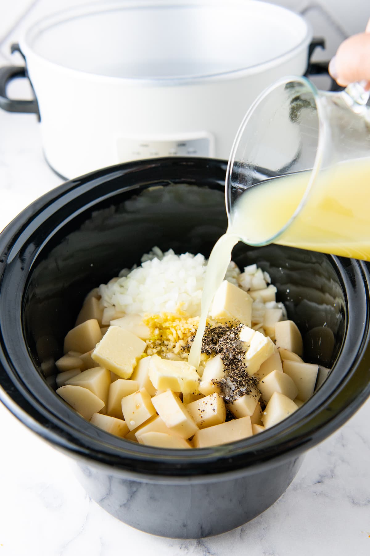 adding broth to a crockpot with potatoes, onions, and seasonings