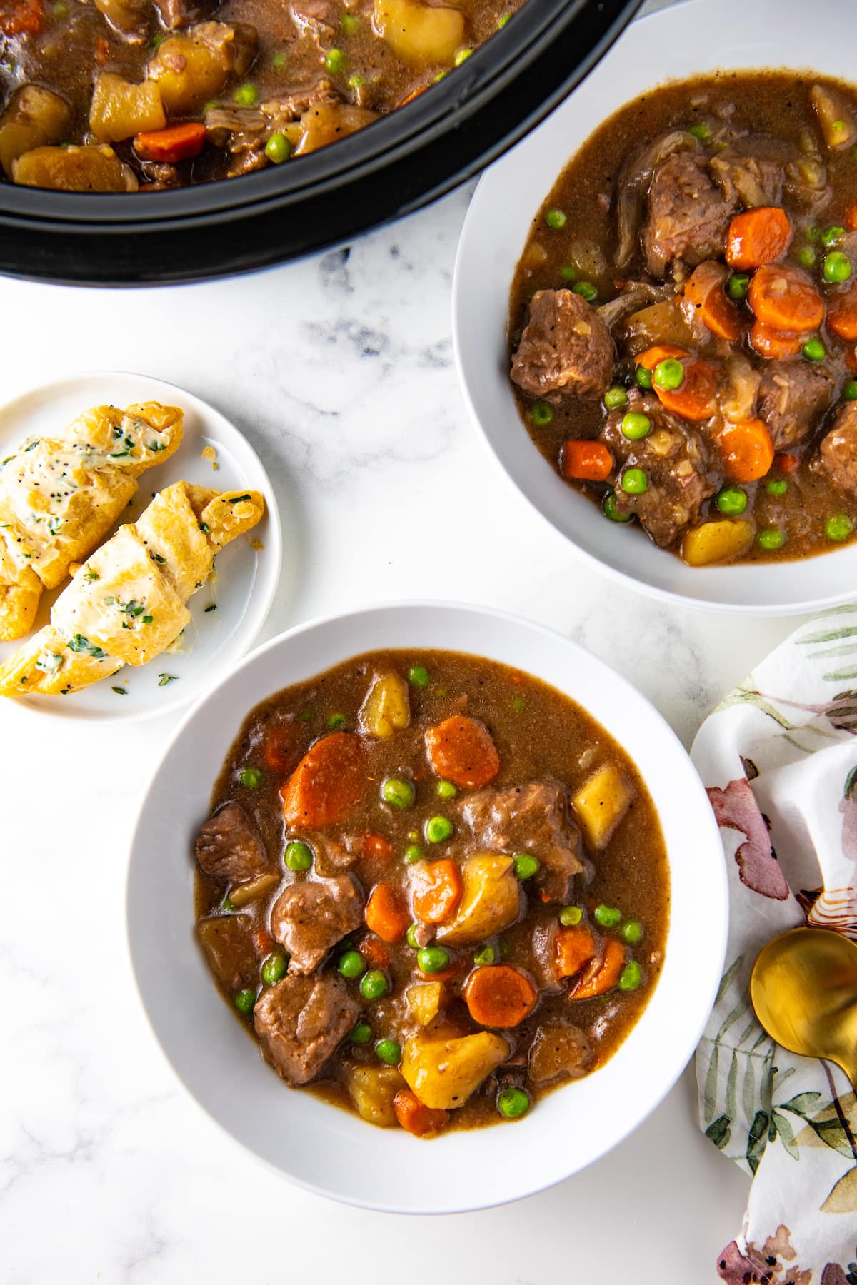 two bowls of beef stew next to a plate with herb crescent rolls