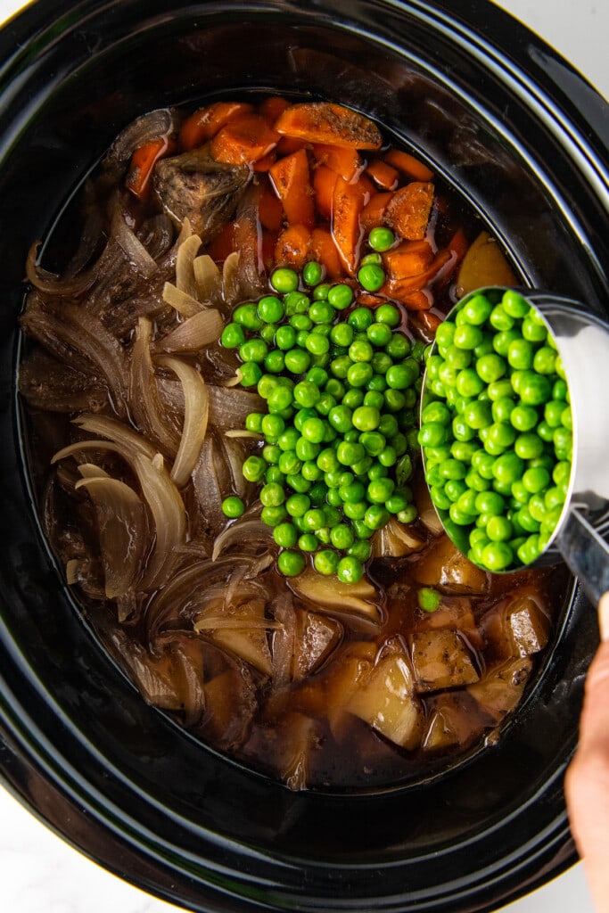 pouring peas into a crock pot with beef, carrots, and potatoes