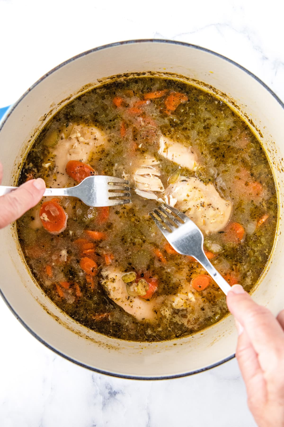 Cooked chicken breasts are being shredded with two forks in a pot of soup.