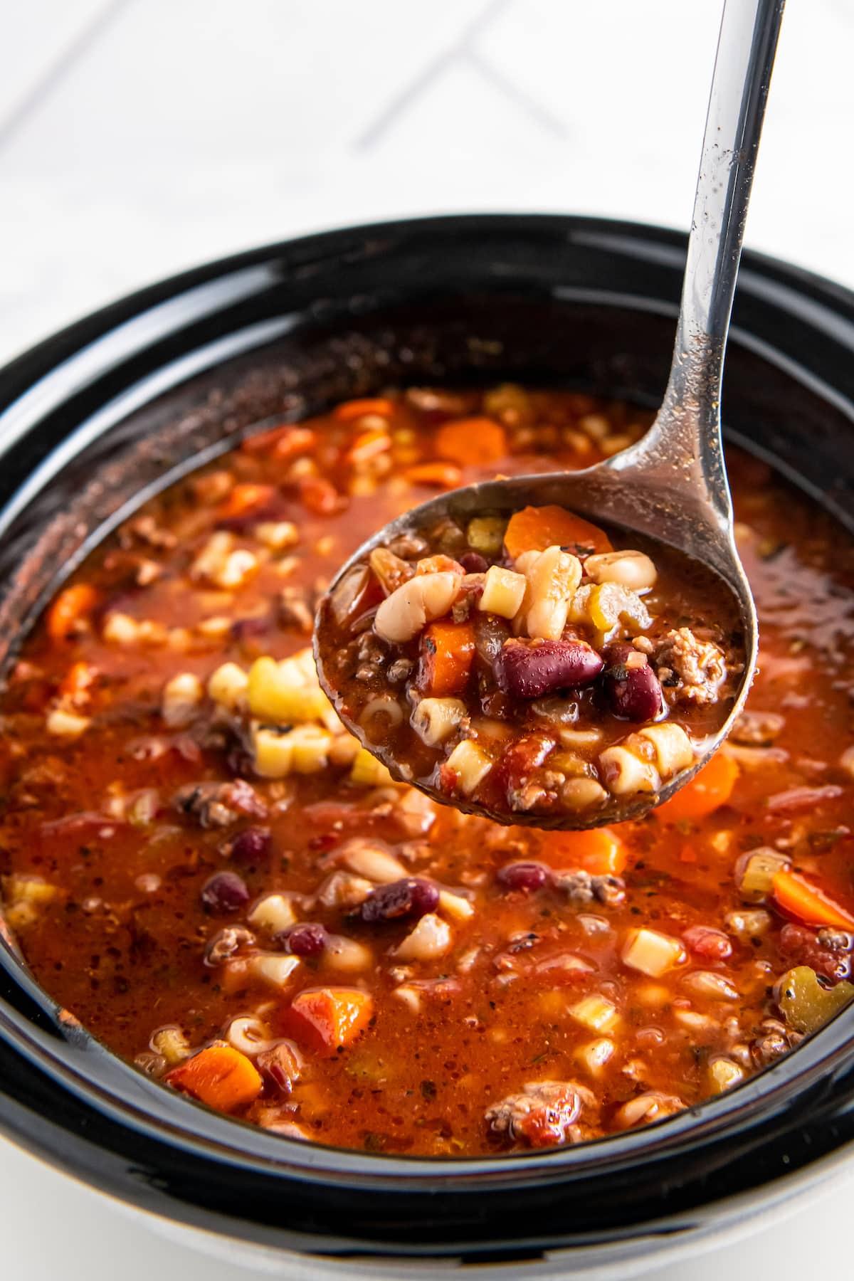 a ladle of soup with a tomato based broth that has ground beef, beans, noodles, and vegetables