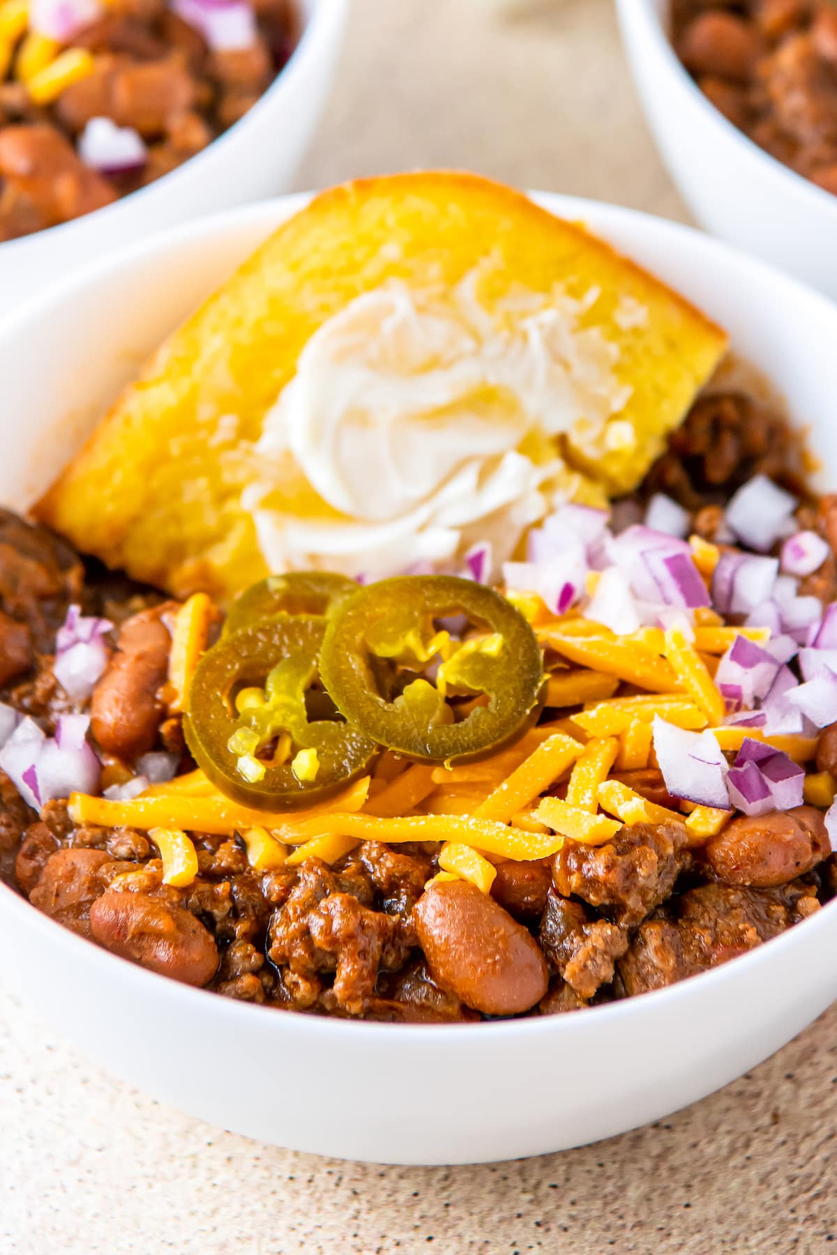 A bowl of beef chili is topped with cheese, onions, cornbread and jalapenos. 