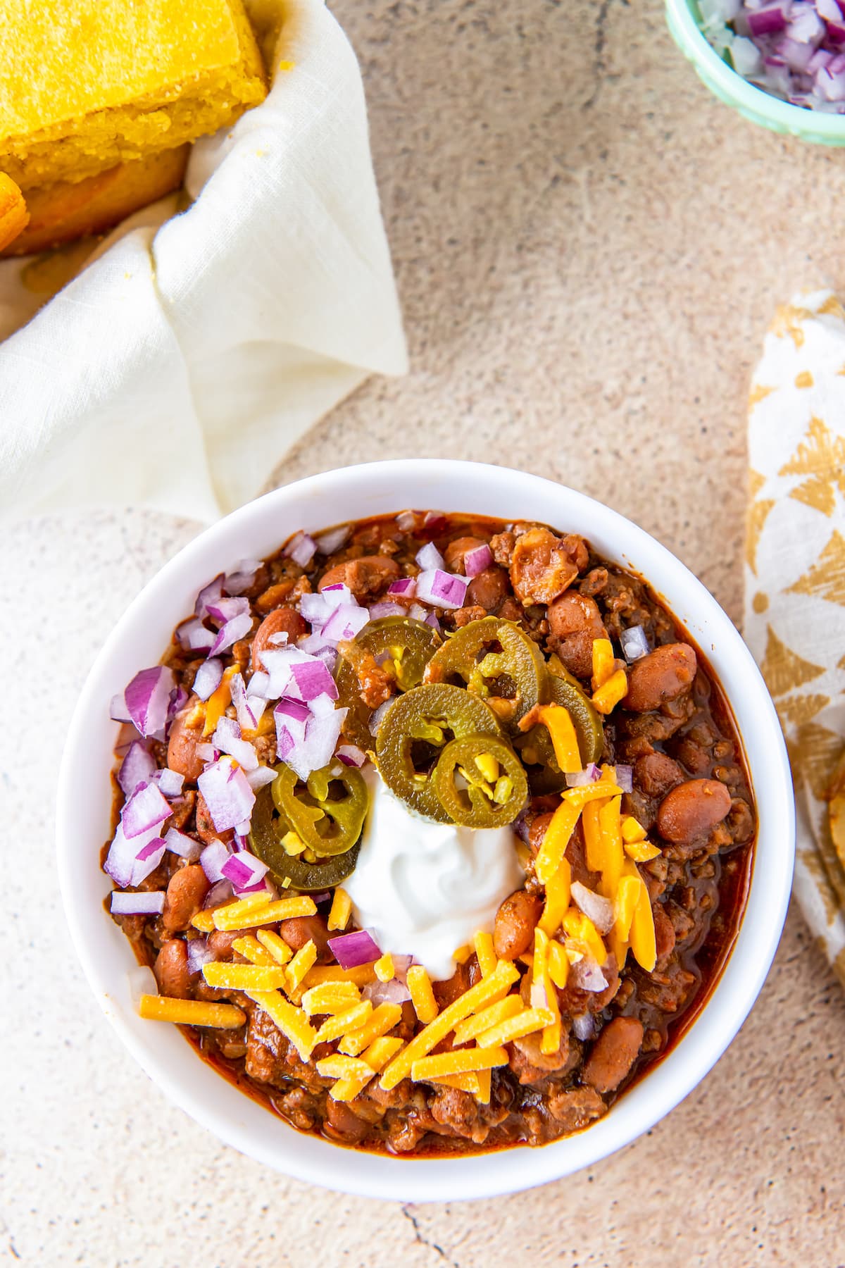 a bowl of chili with toppings like cheese, sour cream, red onion, and pickled jalapenos
