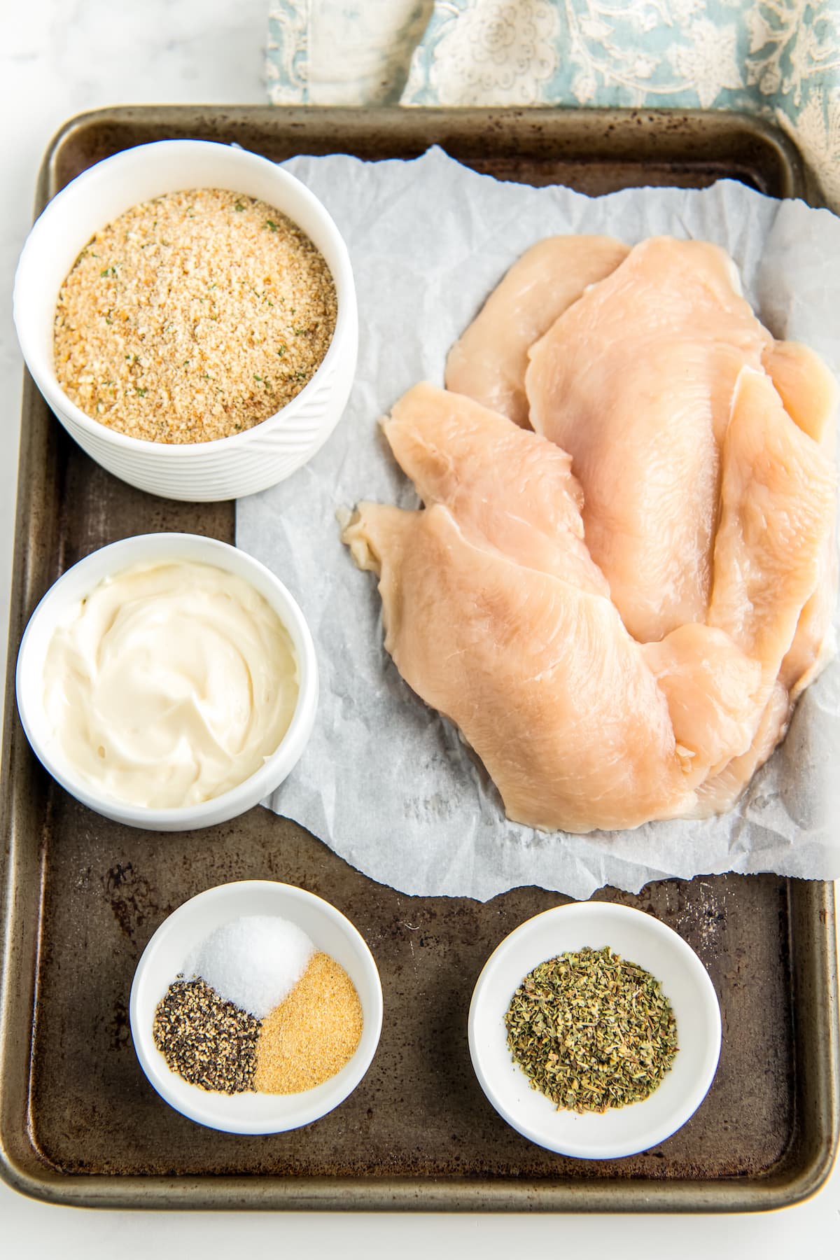 a sheet tray with raw chicken and small bowls of breading, mayonnaise, and spices