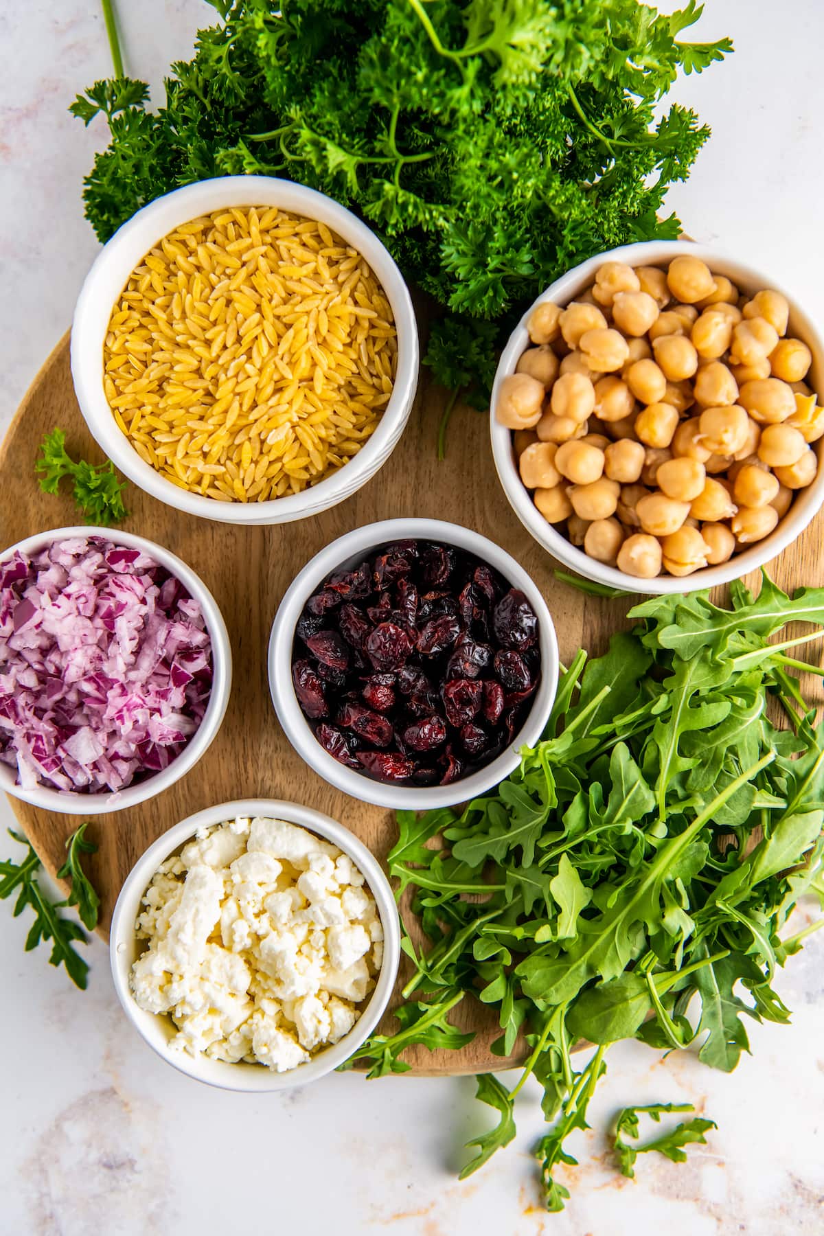 a round cutting board with small bowls that has dried orzo, chickpeas, red onions, dried cranberries, feta, and arugula