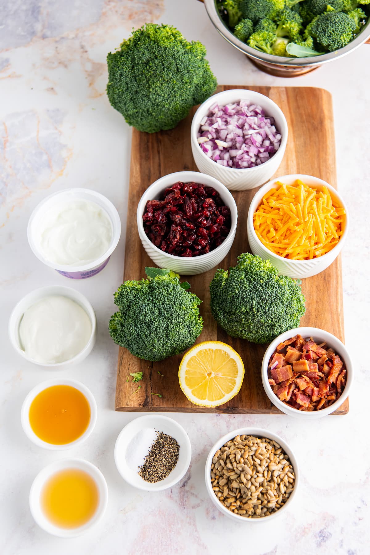 top view of a display of broccoli, seeds, cranberries, and bacon in seperate bowls on a wooden cutting board