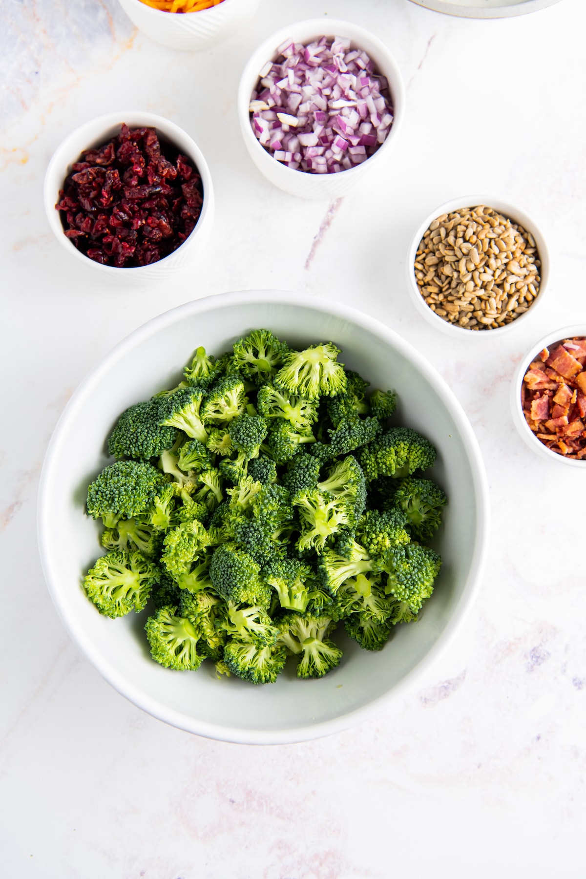 top view of a display of broccoli, seeds, cranberries, and bacon in seperate bowls on a marble countertop