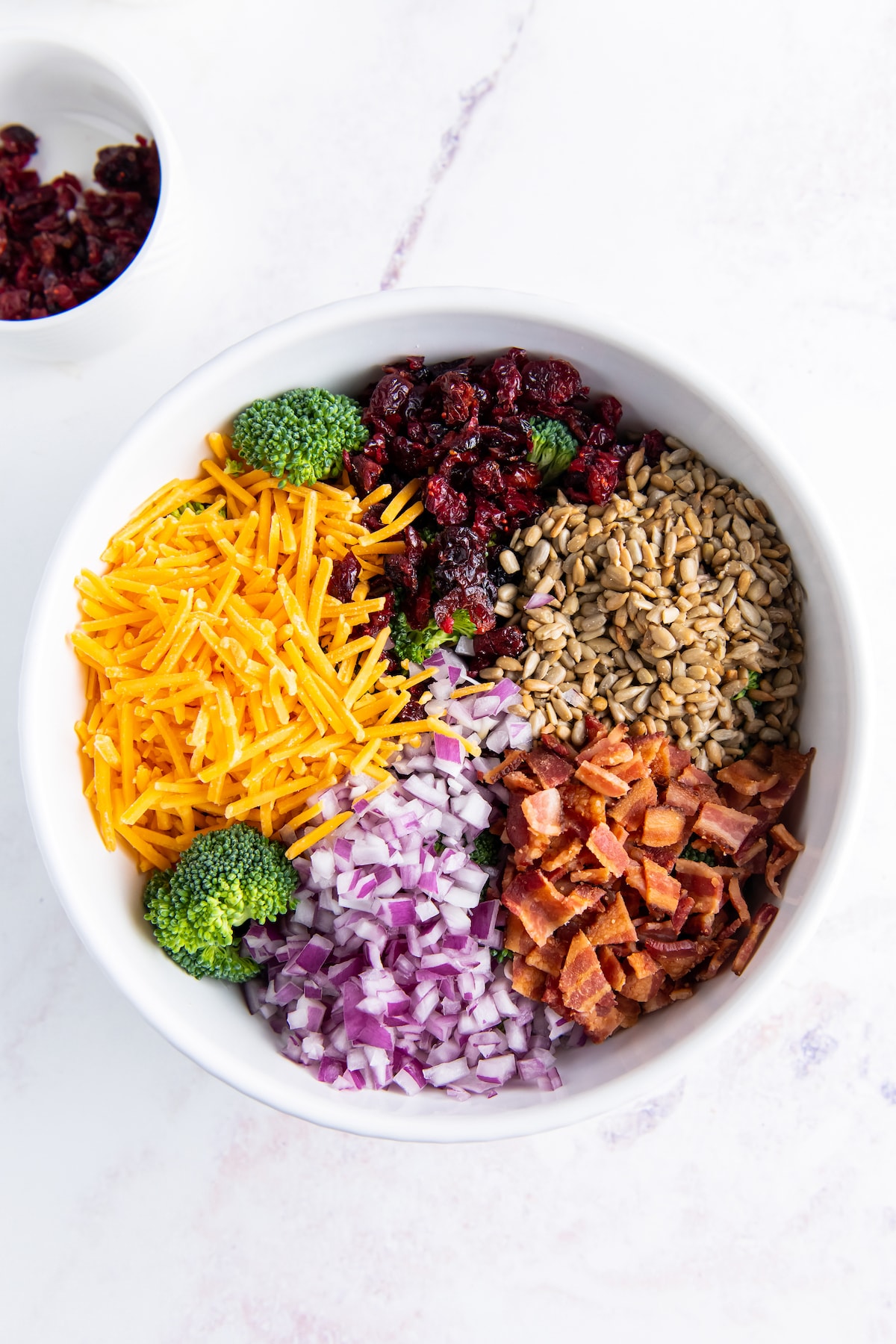 a bowl with seperated ingredients including red onion, bacon, sunflower seeds, broccoli, and shredded cheese