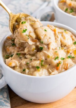 a bowl of chicken with vegetables and dumplings in a small white serving crock