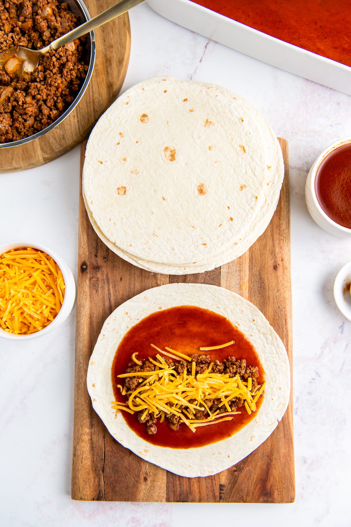 a cutting board with a stack of tortillas being used to roll enchiladas with beef and cheese filling