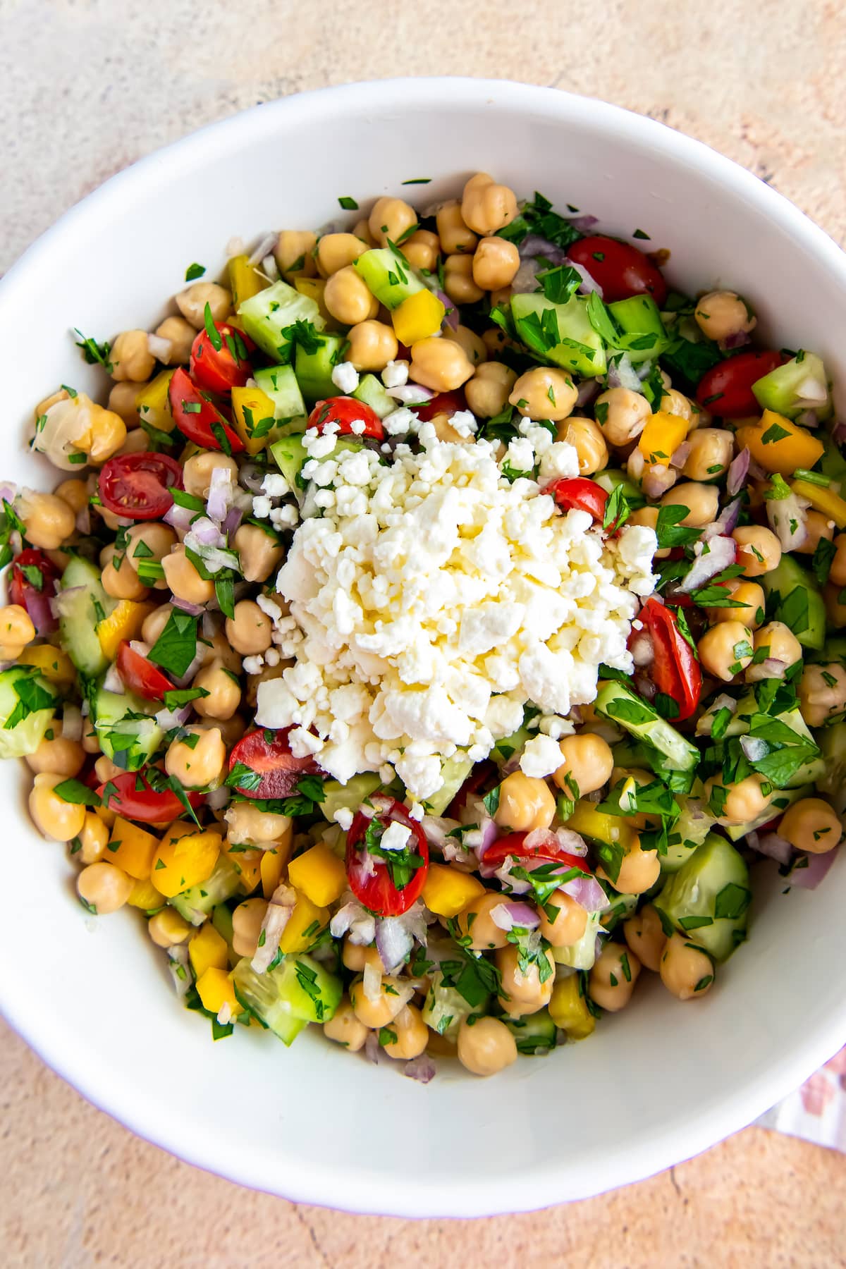 a pile of feta on top of a bowl of salad with chickpeas, cucumbers, tomatoes, and herbs