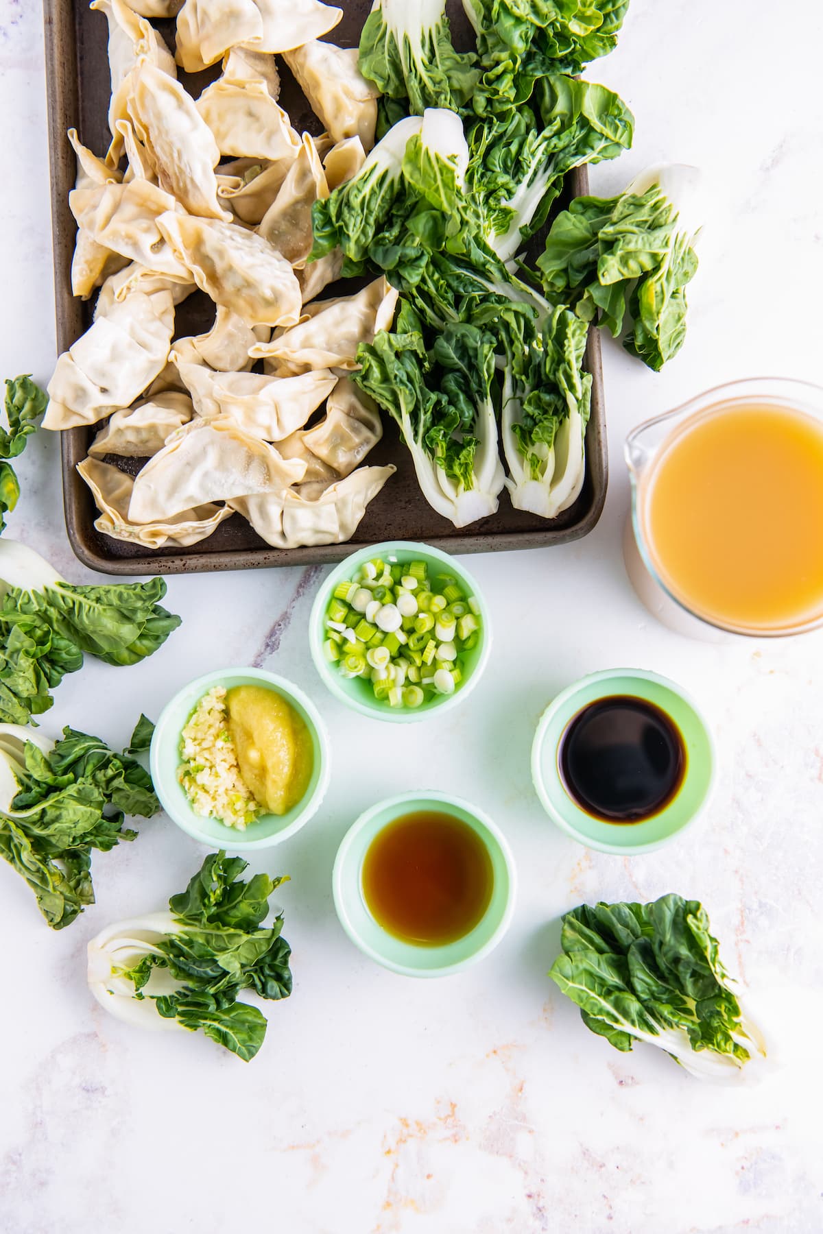an assortment of ingredients to make potsticker soup like small bowls with sauces, oils, bok choy, frozen dumplings and broth