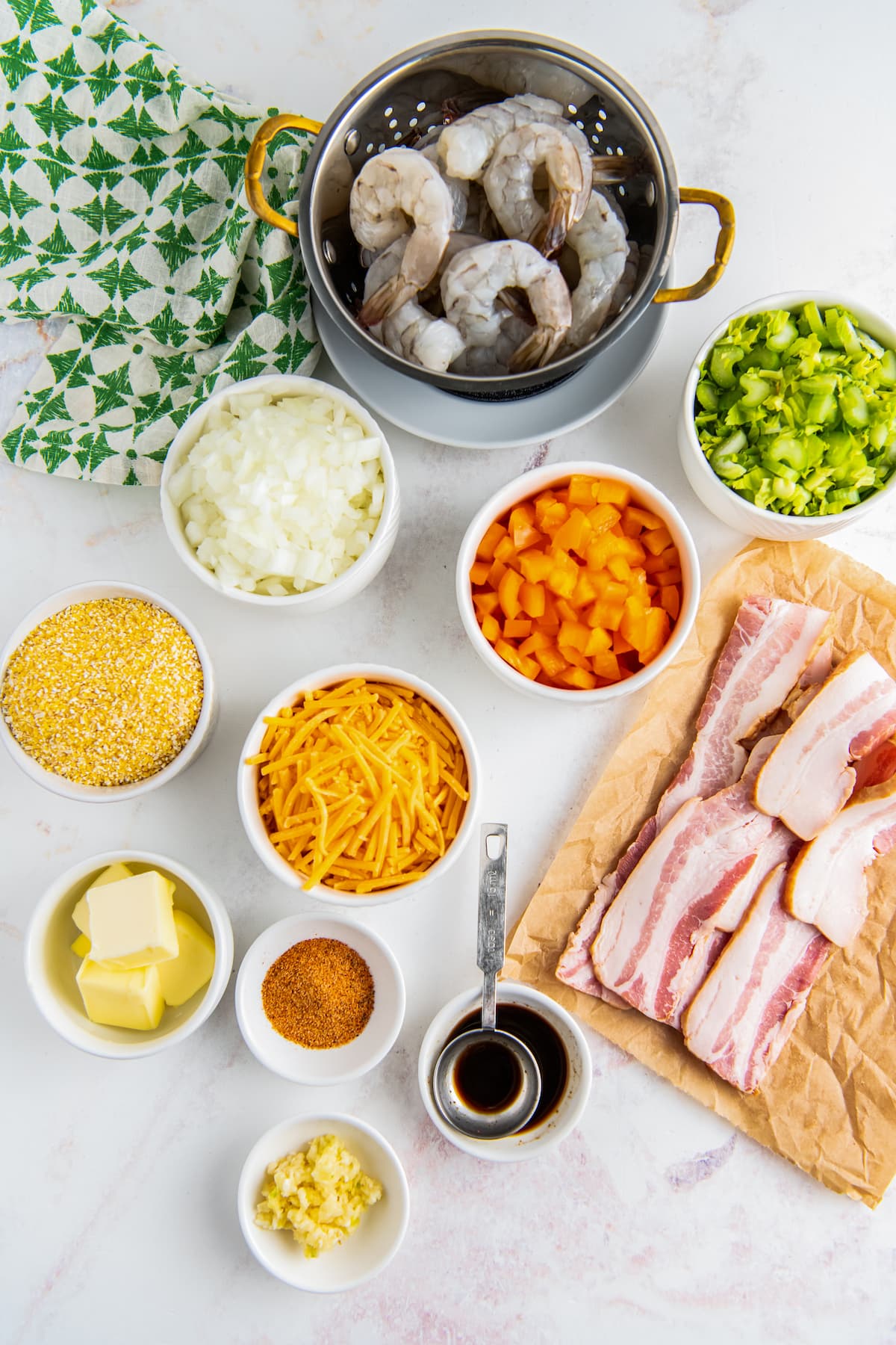 an assortment of ingredients on a marble countertop including bacon, cheese, diced vegetables, butter, seasonings, and shrimp in seperate bowls