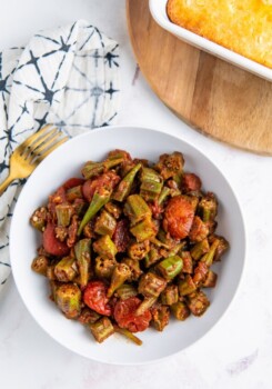 a bowl of stewed okra and tomatoes on a table with a napkin, fork, and wooden cutting board