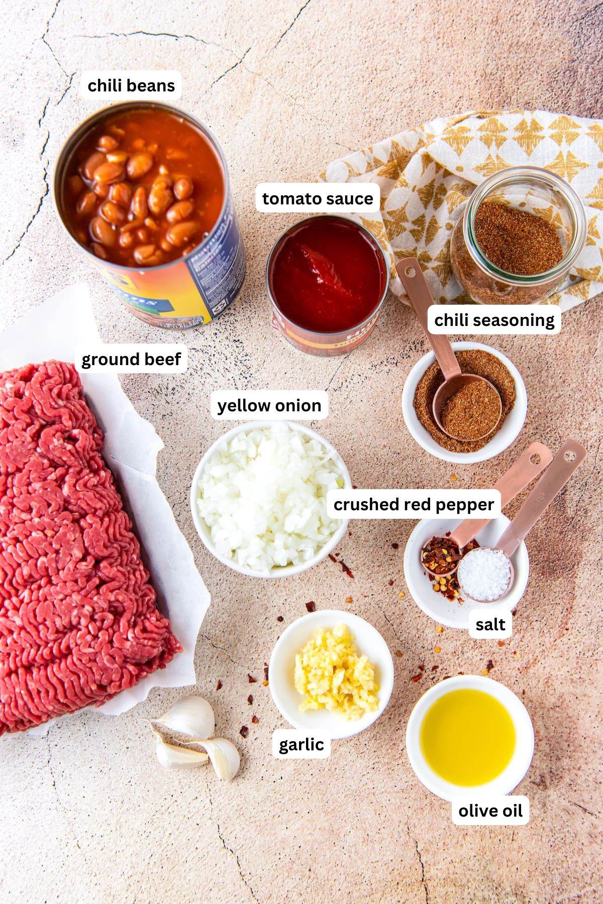 The ingredients for easy chili recipe in order from top to bottom: chili beans, tomato sauce, chili seasoning, ground beef, yellow onion, crushed red pepper, salt, garlic and olive oil.