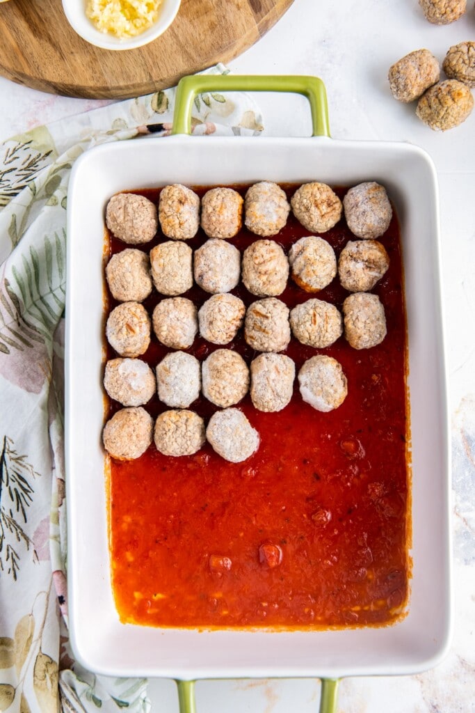 Frozen meatballs on top of a layer of marinara sauce in a baking dish.