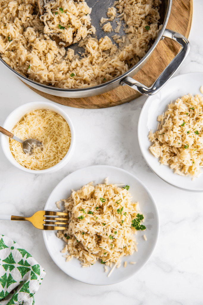 A plate of fluffy and creamy parmesan rice is topped with more shredded cheese and fresh herbs with a skillet filled with rice in the background.