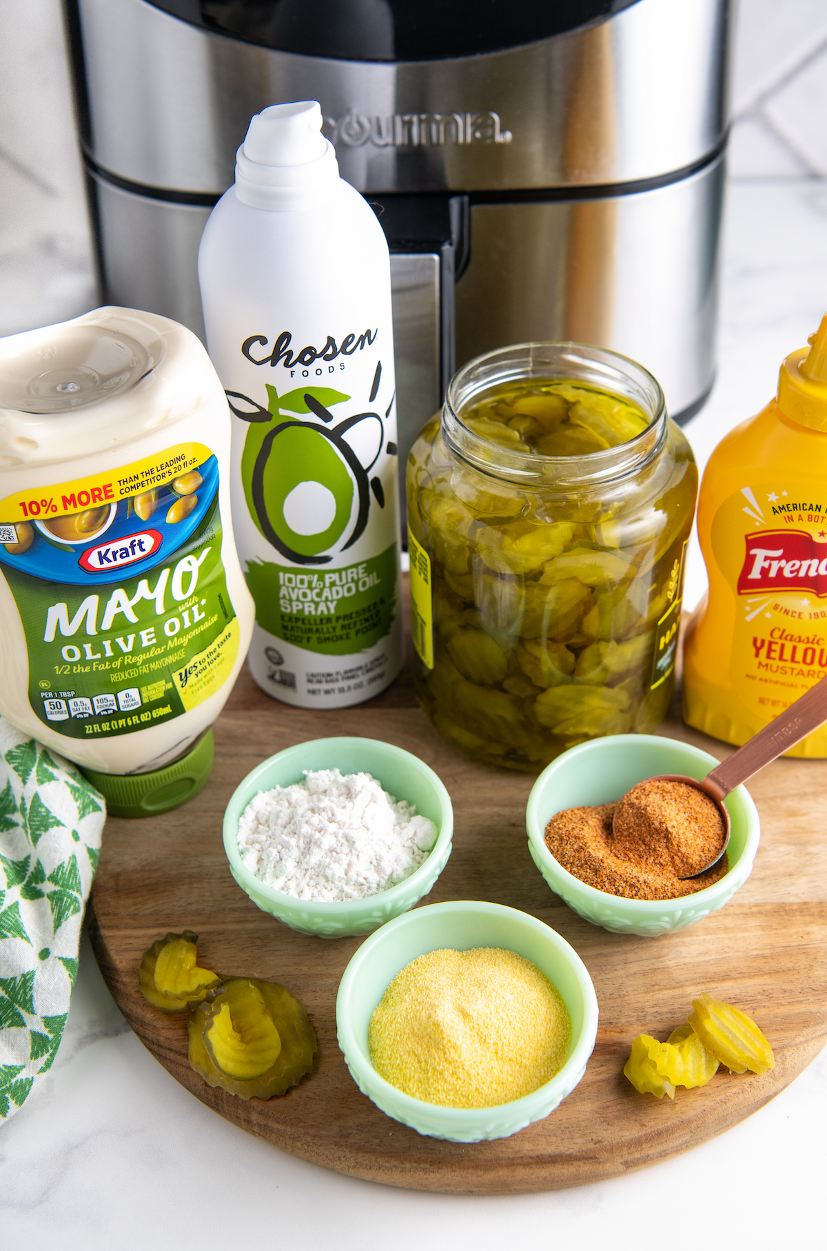 All the ingredients needed for air fryer fried pickles: a jar of pickles, a bottle of oil spray, a bottle of mustard, a bottle of mayo, a bowl of cornmeal, a bowl of flour, and a bowl of cajun seasoning