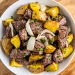 Air fryer steak bites and potatoes with onions in a bowl with fresh herbs on top.