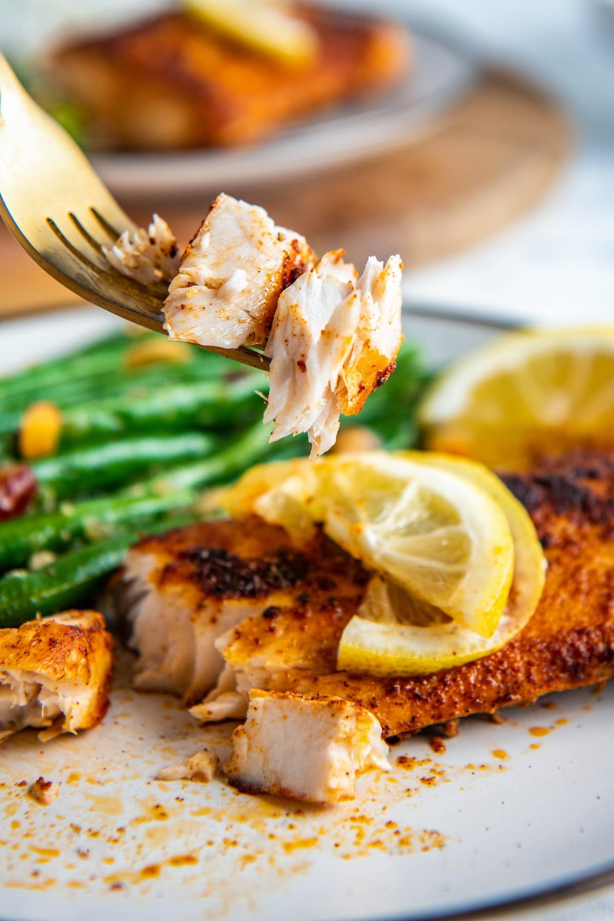 A fork with fish on it, with a piece of blackened mahi mahi and green beans in the background