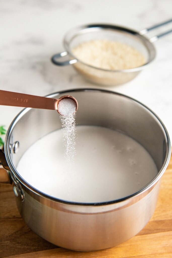 A measuring spoon pouring salt into a pot of water and coconut milk, with a bowl of uncooked rice in the background