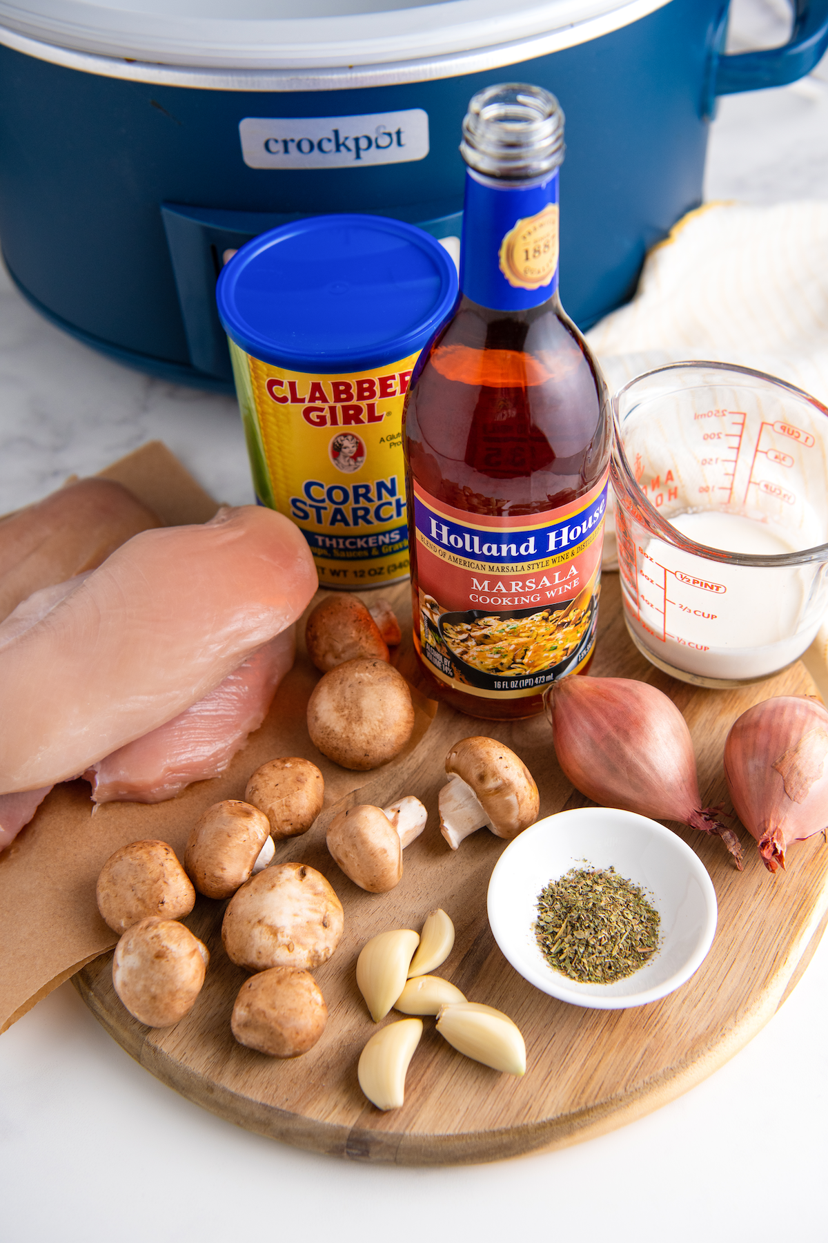 The ingredients for chicken marsala: a pile of raw chicken breasts, a bunch of mushrooms, two shallots, a bowl of Italian seasoning, a pyrex of cream, a bottle of marsala wine, and a tin of corn starch