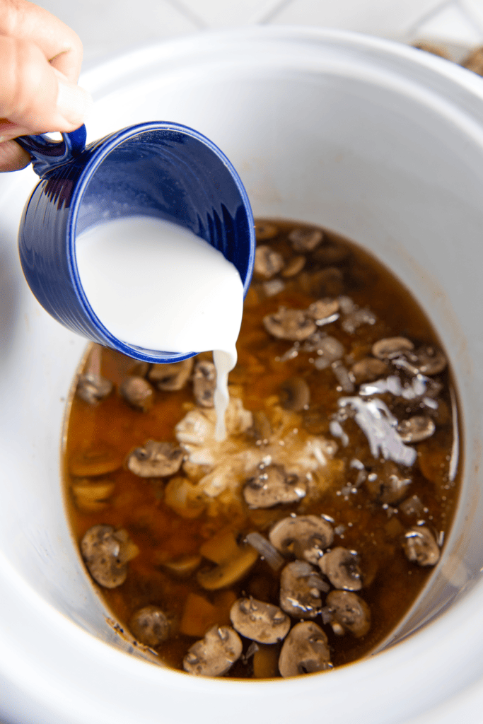 A measuring cup pouring a slurry into a crock pot full of broth and mushrooms