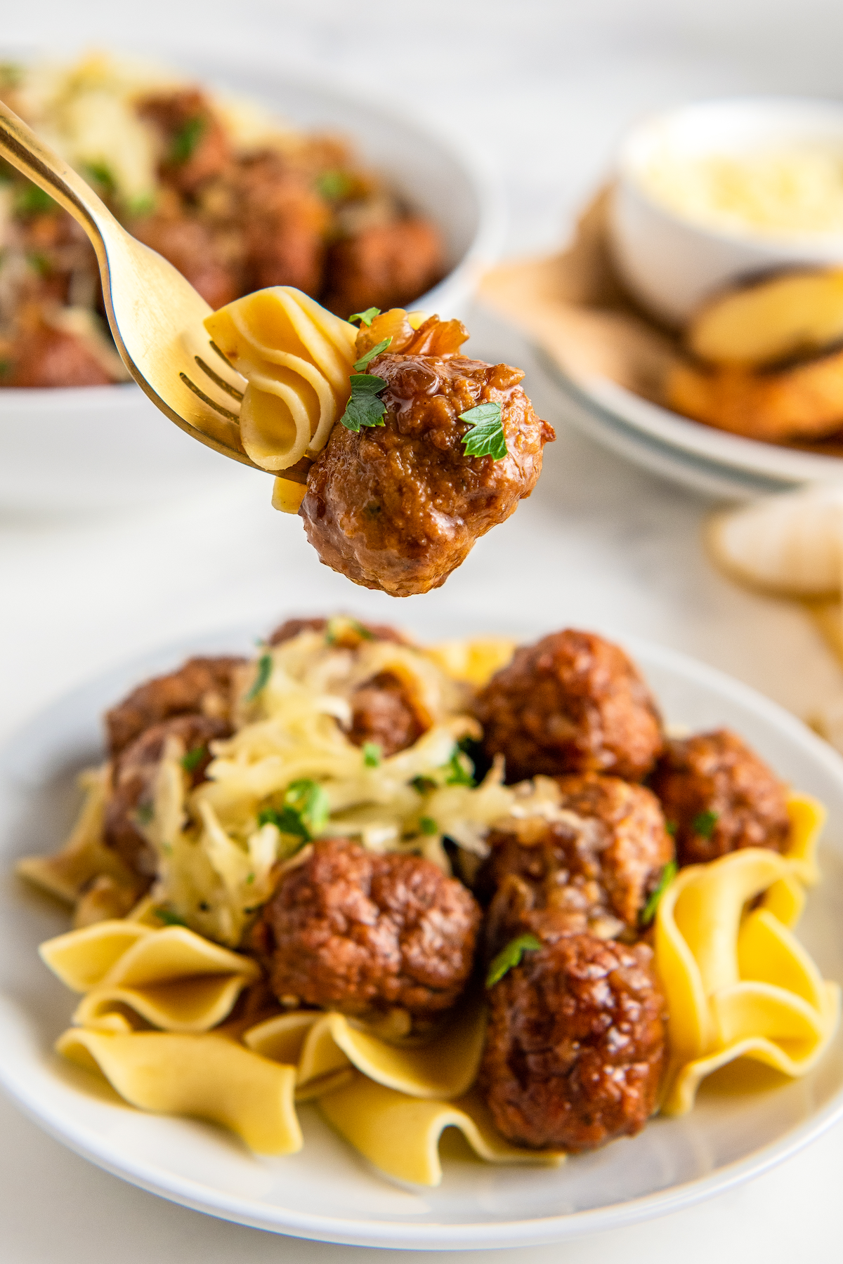 A meatball and some pasta on a fork, above a plate of pasta and meatballs, with another plate in the background