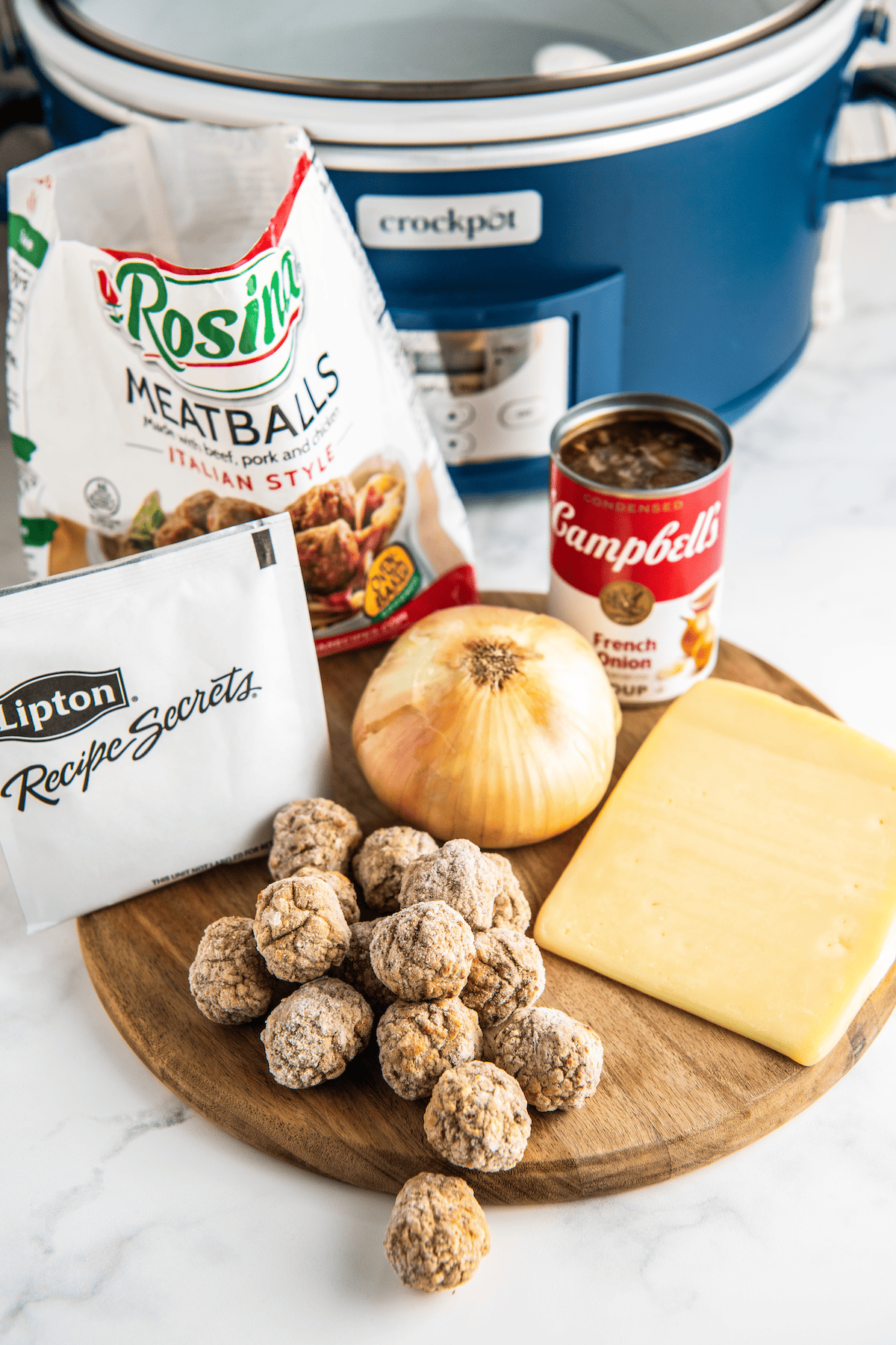 The ingredients needed for French onion meatballs: a bag of frozen meatballs, a pile of frozen meatballs, a packet of Lipton onion soup mix, a can of Campbells French onion soup, a yellow onion, and a block of gruyere cheese, in front of a crockpot