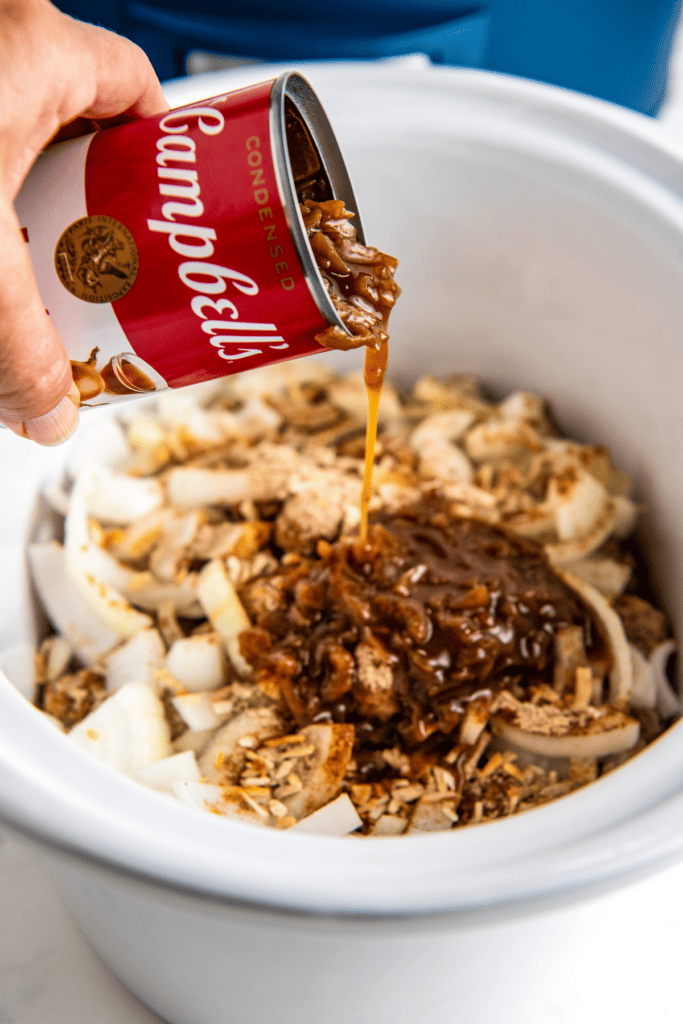 A can of Campbells French onion soup being poured into a crockpot full of meatballs, onions, and seasoning