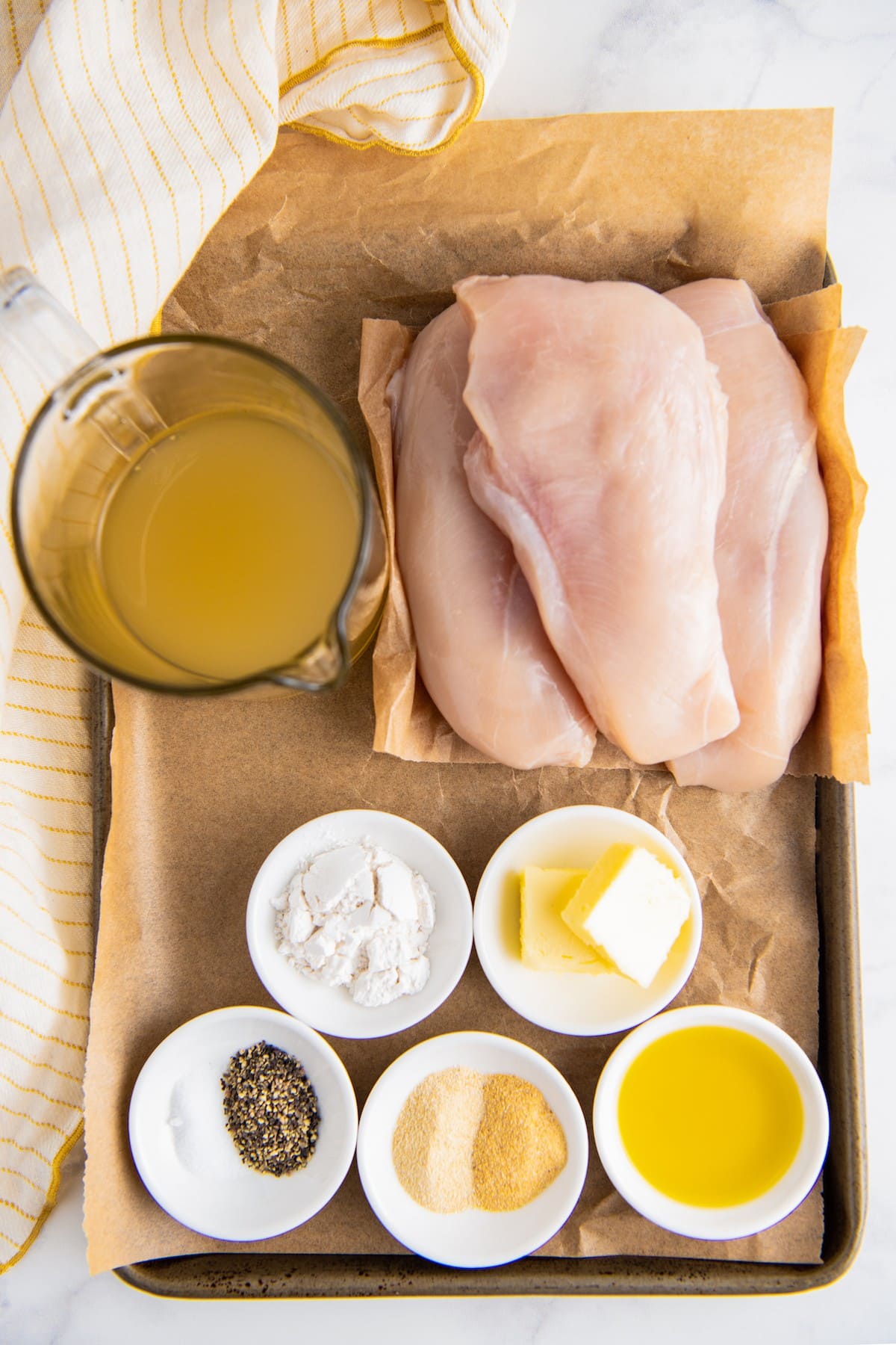 Overhead view of the ingredients needed to make chicken and gravy: a pile of raw chicken breasts, a pyrex of chicken broth, a bowl of flour, a bowl of butter, a bowl of oil, a bowl of salt and pepper, and a bowl of granulated garlic and onion powder
