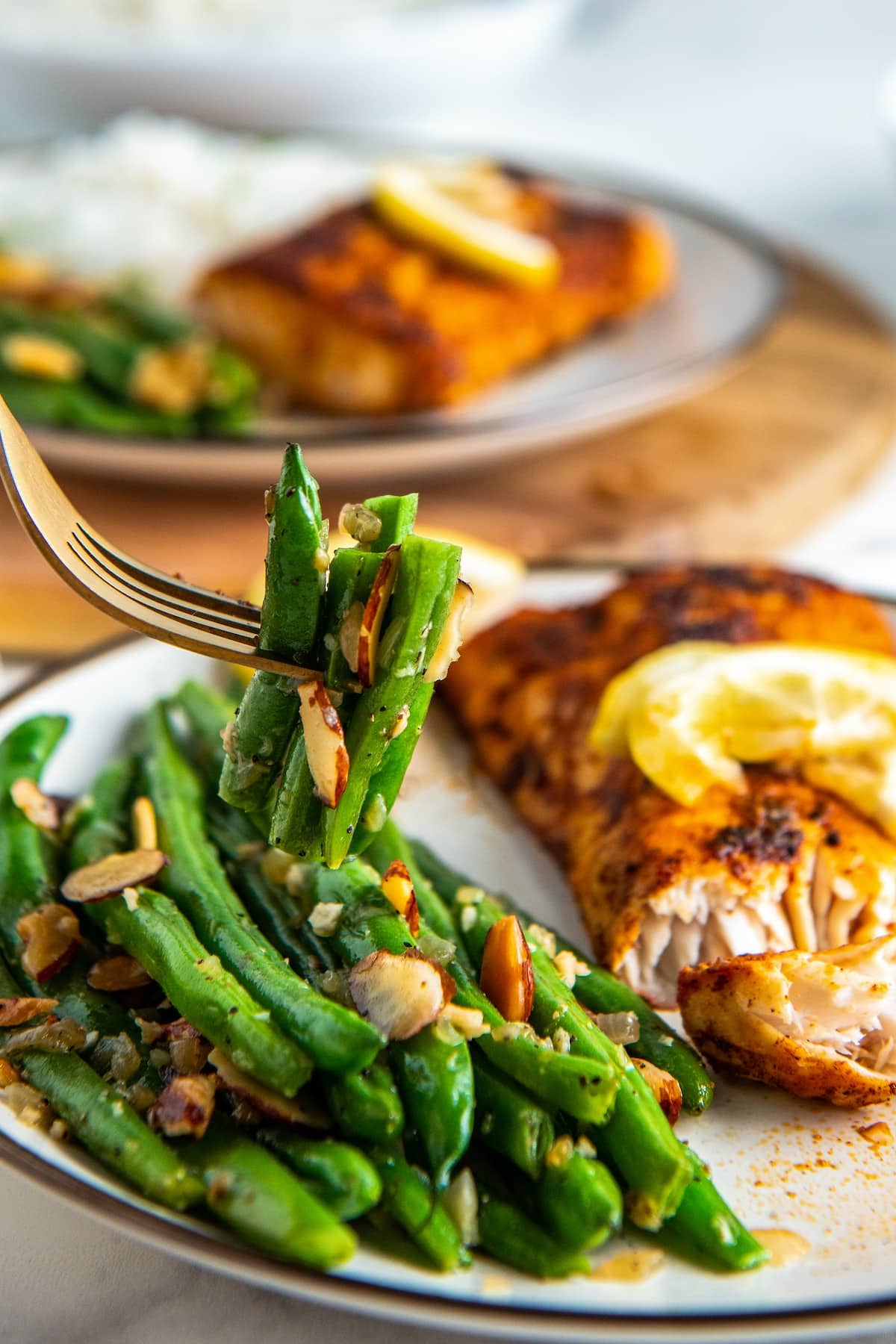 A fork with green beans almondine on it, with two plates in the background filled with green beans and salmon