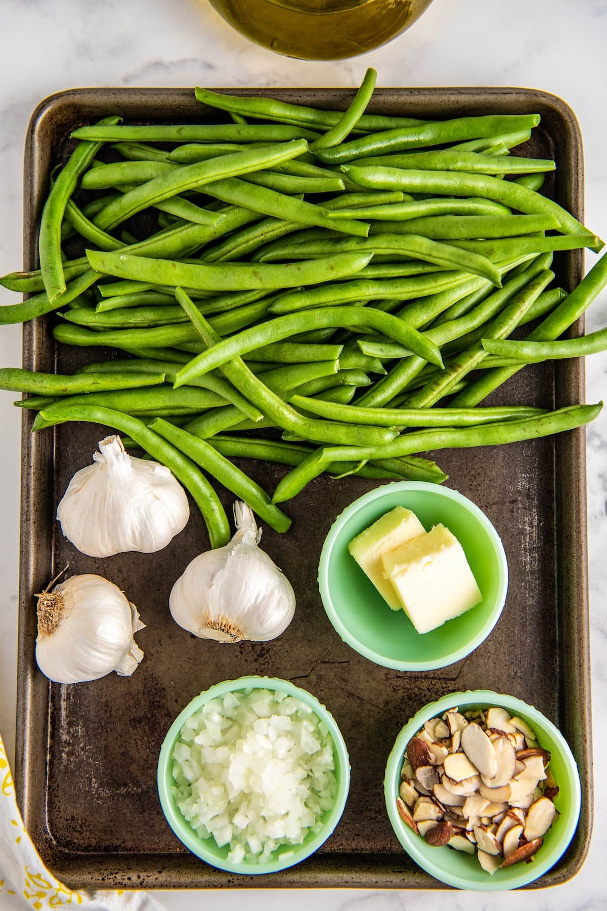 Overhead view of the ingredients needed for green beans almondine: raw green beans, three heads of garlic, a bowl of butter, a bowl of sliced almonds, and a bowl of diced shallot