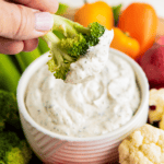 A hand dipping a piece of broccoli into a bowl of ranch, surrounded by carrots and peppers