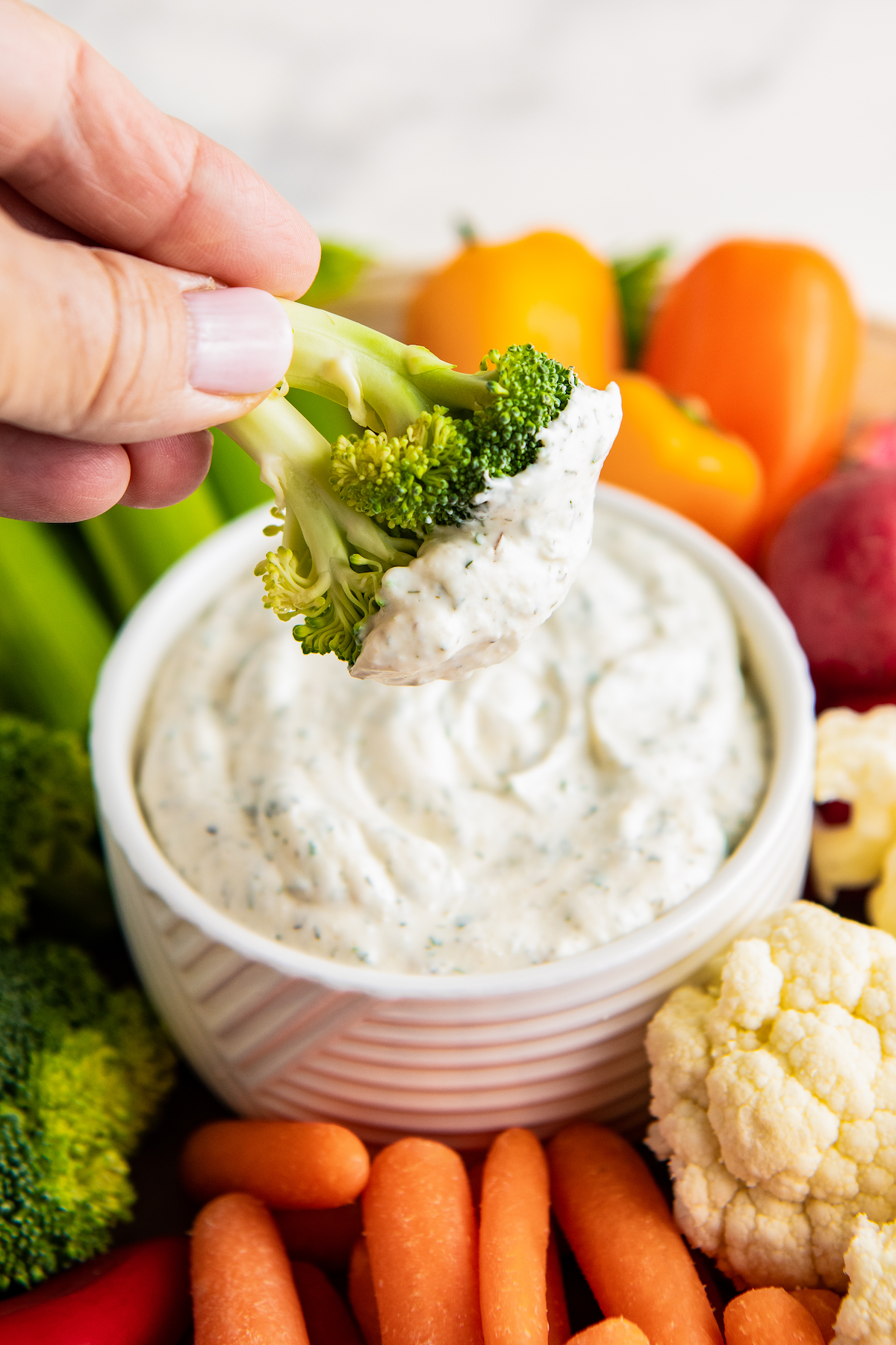 A hand dipping a piece of broccoli into a bowl of ranch, surrounded by carrots and peppers