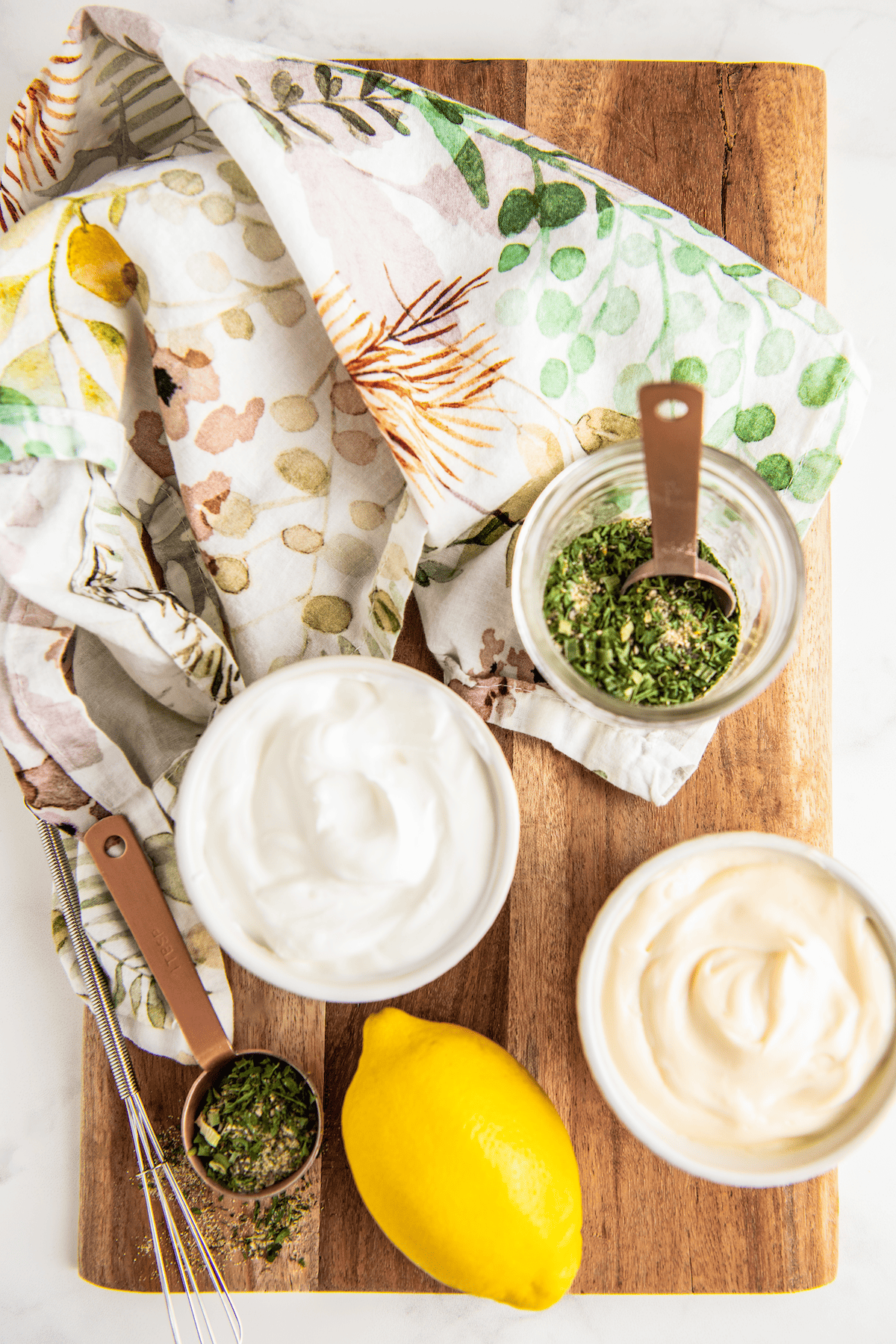 Overhead view of the ingredients needed for ranch dressing: a bowl of sour cream, a bowl of mayo, a lemon, and a jar of ranch seasoning with a spoon in it, next to a measuring spoon and kitchen towels