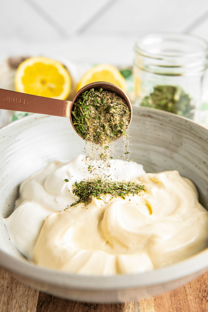 A measuring spoon pouring ranch seasoning into a bowl of mayo and sour cream, with lemons in the background