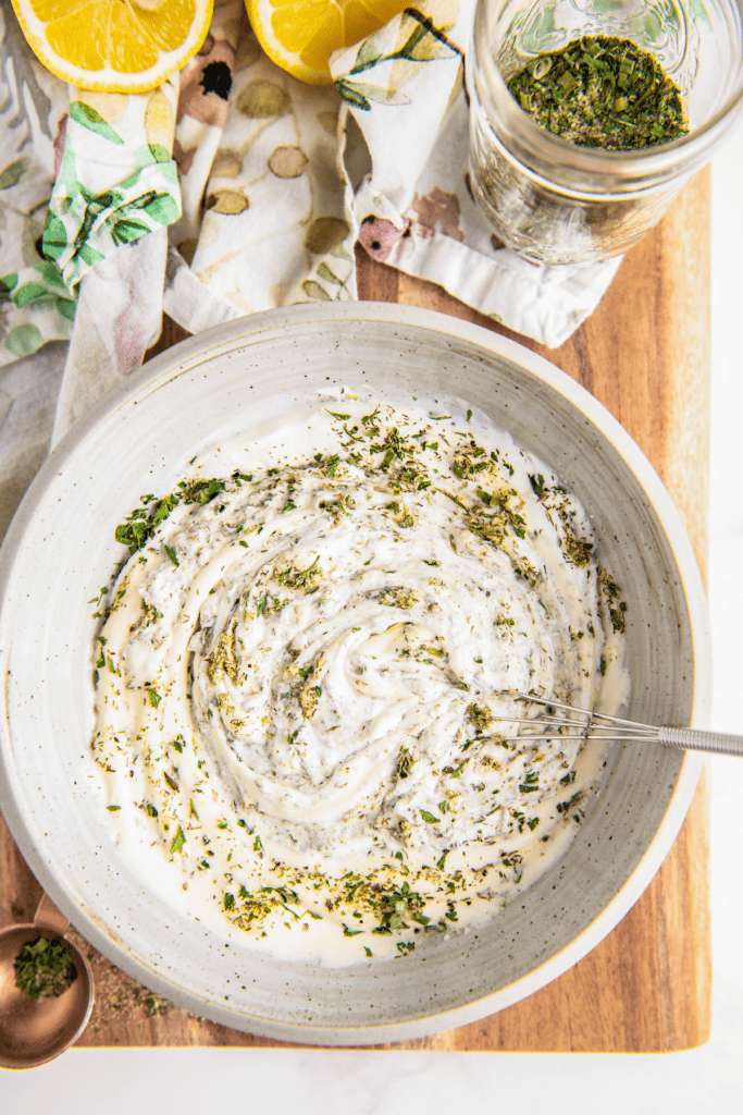 A bowl of partially mixed ranch dressing, with i whisk in it, next to a jar of ranch seasoning and a lemon