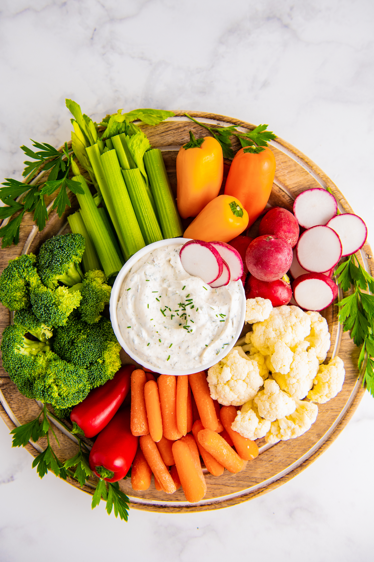 Overhead view of a round tray filled with celery, broccoli, carrots, peppers, radishes, and cauliflower, with a bowl of ranch in the middle